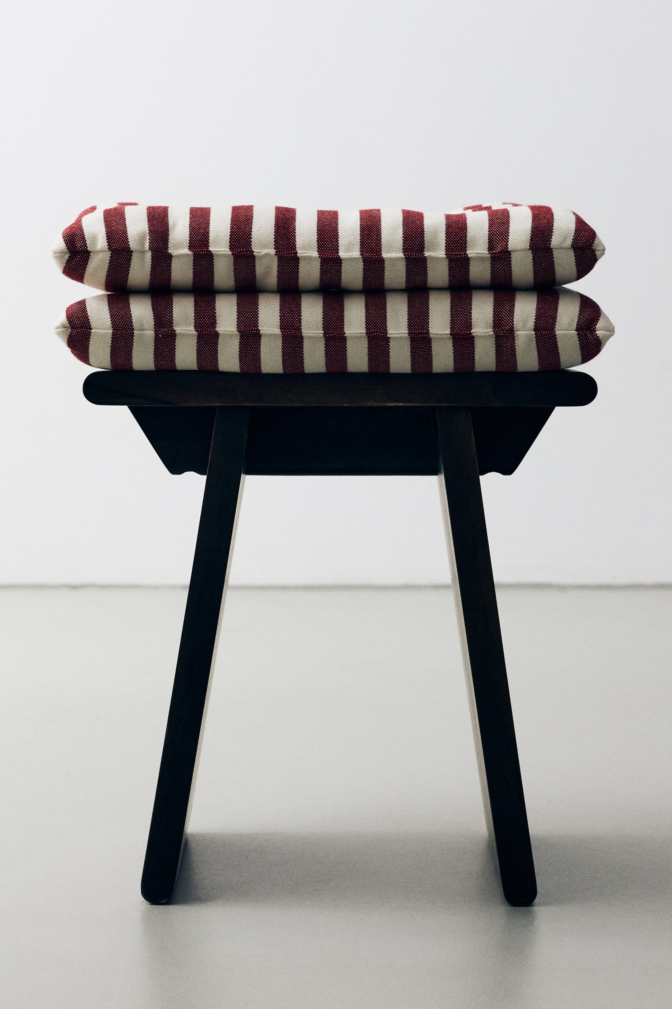 View larger image: Two padded seat cushions with vertical red and off-white stripes are stacked on a dark wooden stool, shown from the front to highlight their plump, rectangular shape. The cushions feature a textured cotton weave and visible thickness.