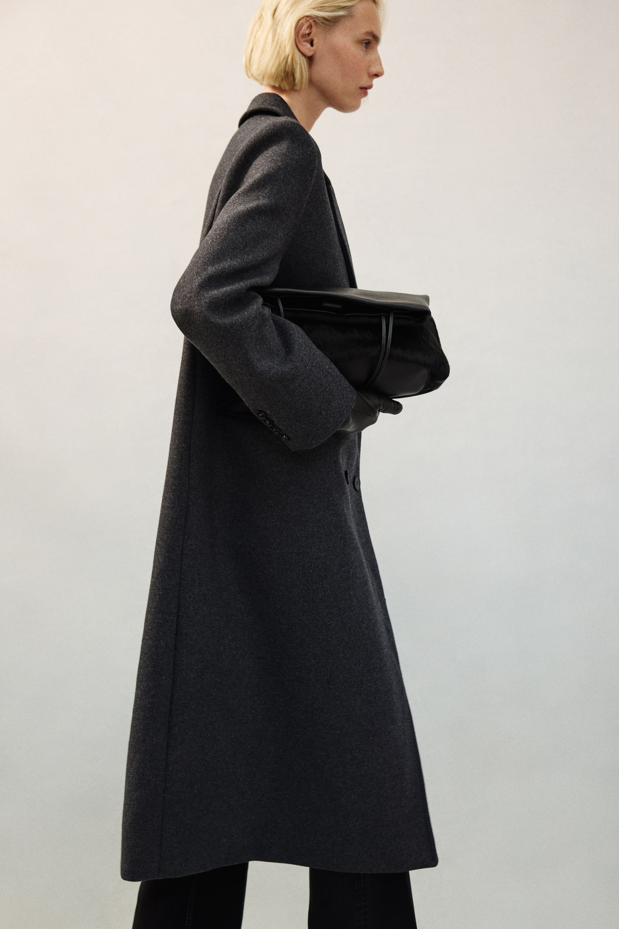 View larger image: A long, dark grey wool-blend coat with structured shoulders, peak lapels, and button cuffs is being worn by the model. The model is carrying a black leather handbag tucked under their arm while wearing black leather gloves.
