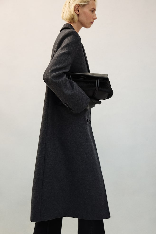 A long, dark grey wool-blend coat with structured shoulders, peak lapels, and button cuffs is being worn by the model. The model is carrying a black leather handbag tucked under their arm while wearing black leather gloves. - 1
