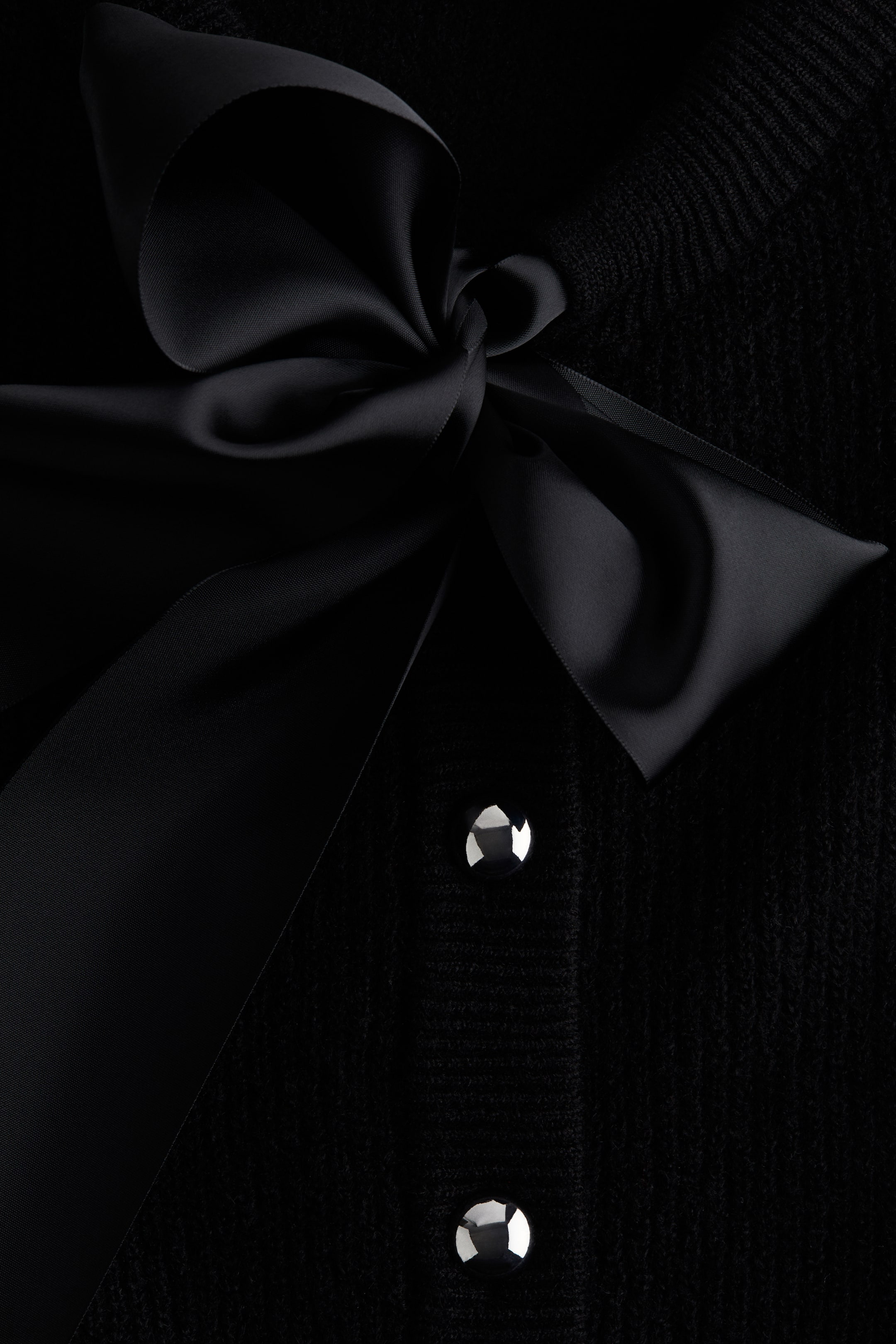 Grotere afbeelding bekijken: Een zwart gebreid vest heeft een grote zwarte satijnen strik die bij de halslijn is vastgebonden. Twee ronde, zilverkleurige gefacetteerde knopen zijn zichtbaar langs de voorkant van de sluiting.