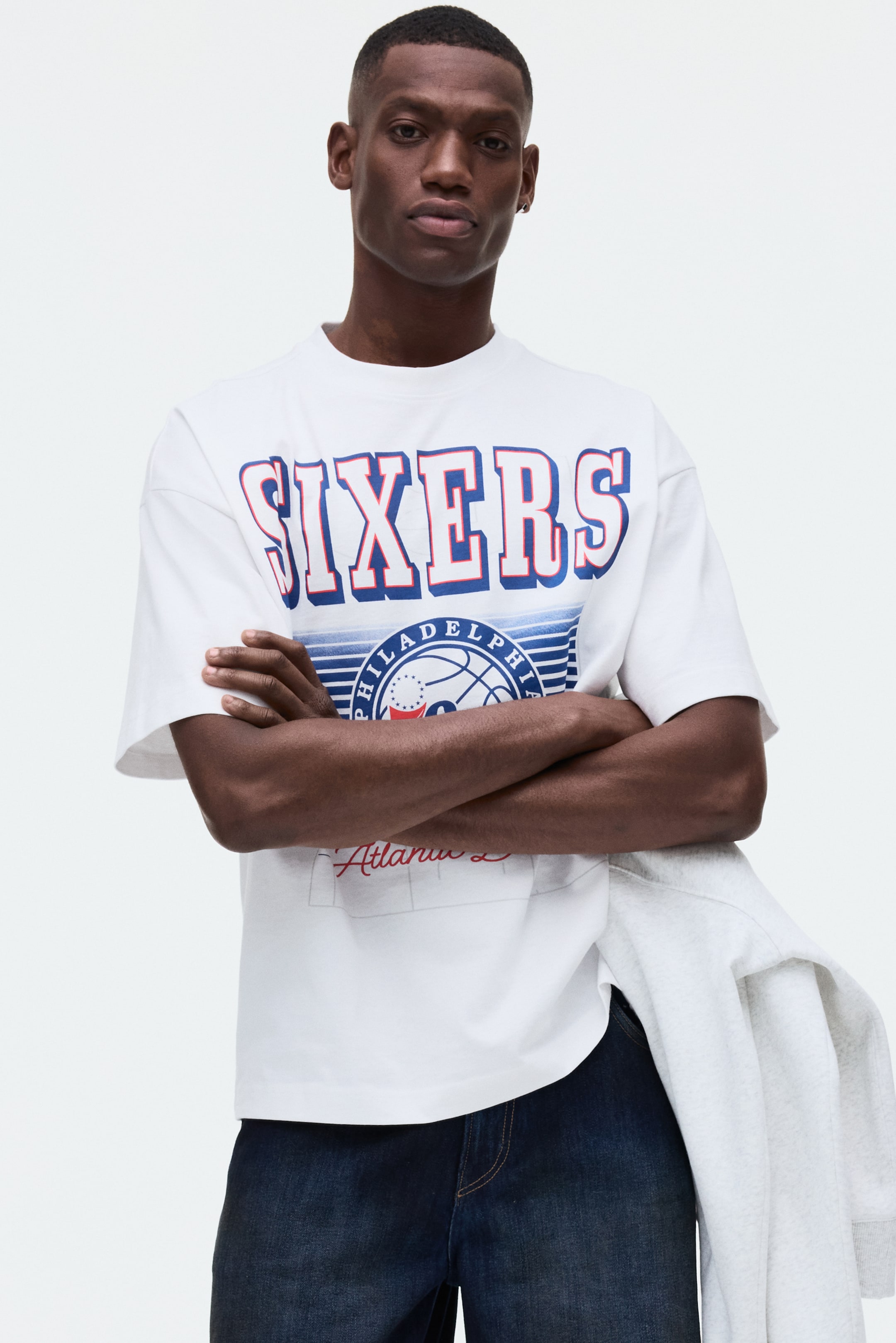 Visa större bild: En vit oversize t-shirt är vänd framåt, med en stor "SIXERS"-grafik på bröstet med blå bokstäver och röda och vita konturer. Nedanför detta syns en cirkulär basketgrafik med texten "PHILADELPHIA", omgiven av blå ränder, och "Atlantic" är skrivet i röd kursiv stil under den.