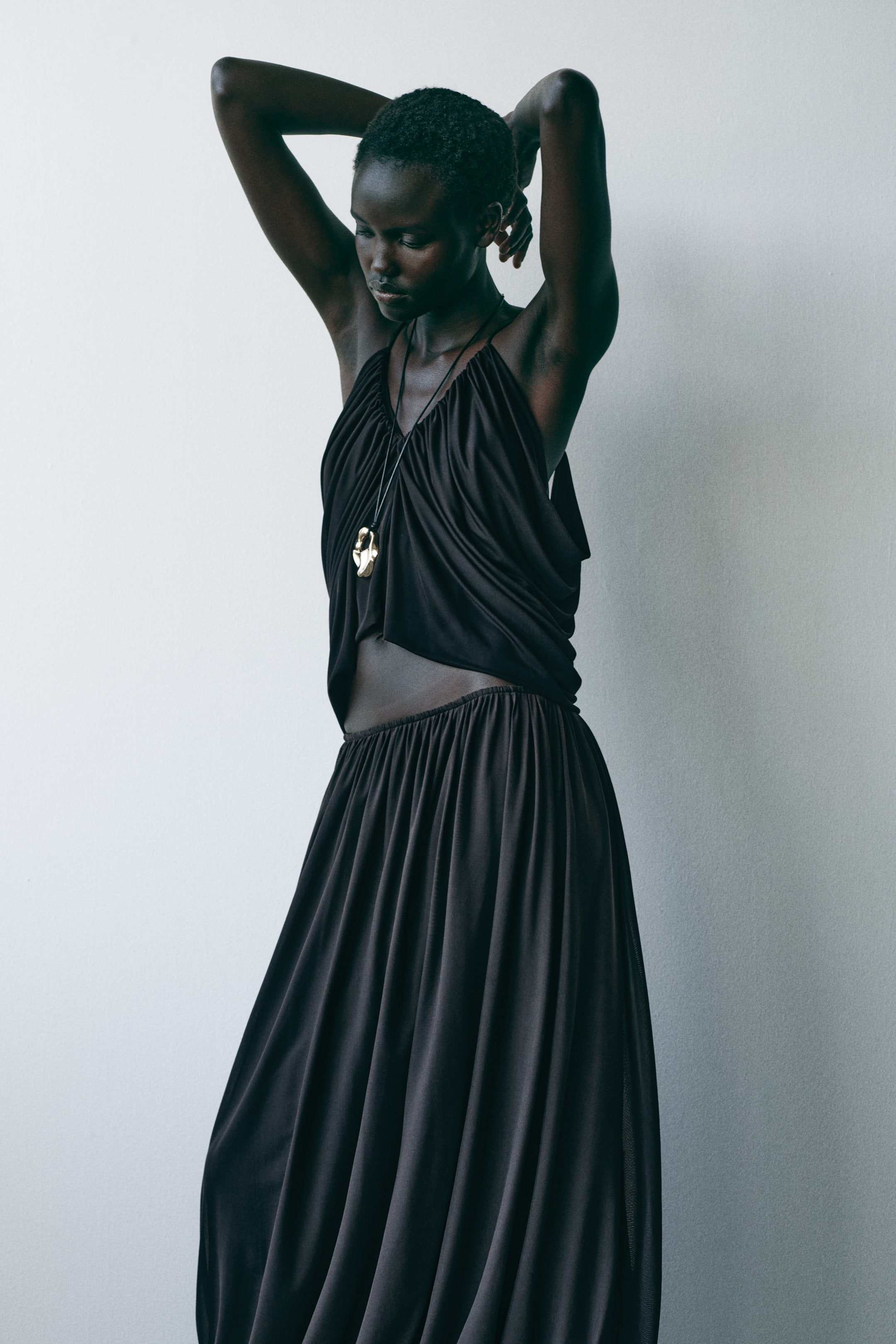 Größeres Bild ansehen: Ein schwarzes zweiteiliges Ensemble präsentiert ein drapiertes Oberteil im Halterneck-Stil mit einem gerafften V-Ausschnitt und einem elastischen, gekürzten Saum, kombiniert mit einem fließenden Maxirock, der einen gerafften Bund und eine volle Silhouette zeigt, aus einem leichten Winkel präsentiert. Eine lange schwarze Kordelkette mit einem goldfarbenen, organisch geformten Anhänger ist über dem Oberteil geschichtet.