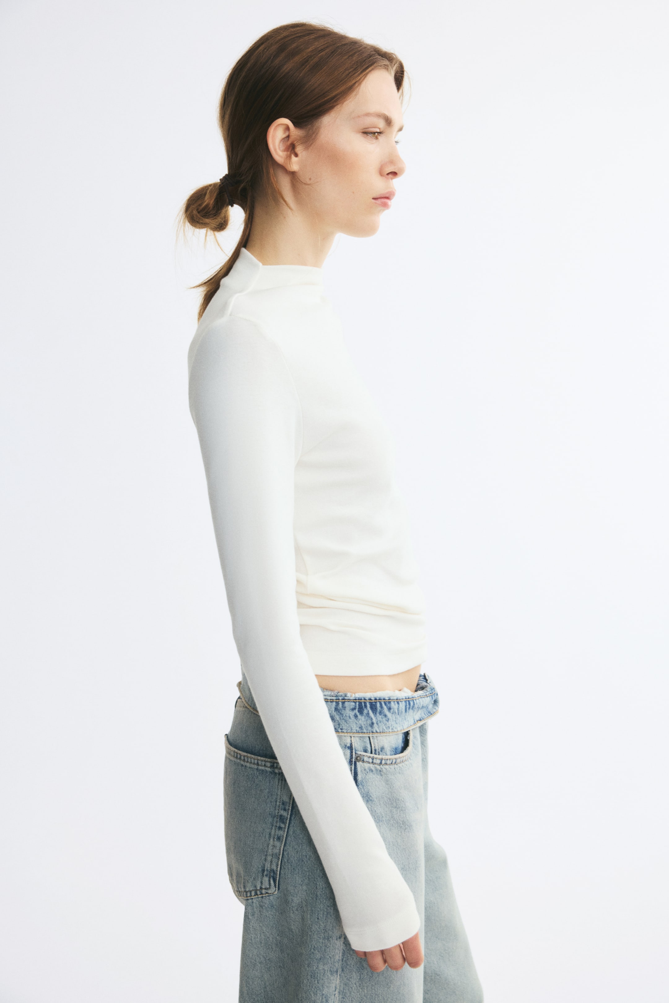 View larger image: The model is wearing a white fitted jersey top with a high neck and long sleeves. Styled with light-wash denim jeans, the top highlights its snug fit along the torso and sleeves.