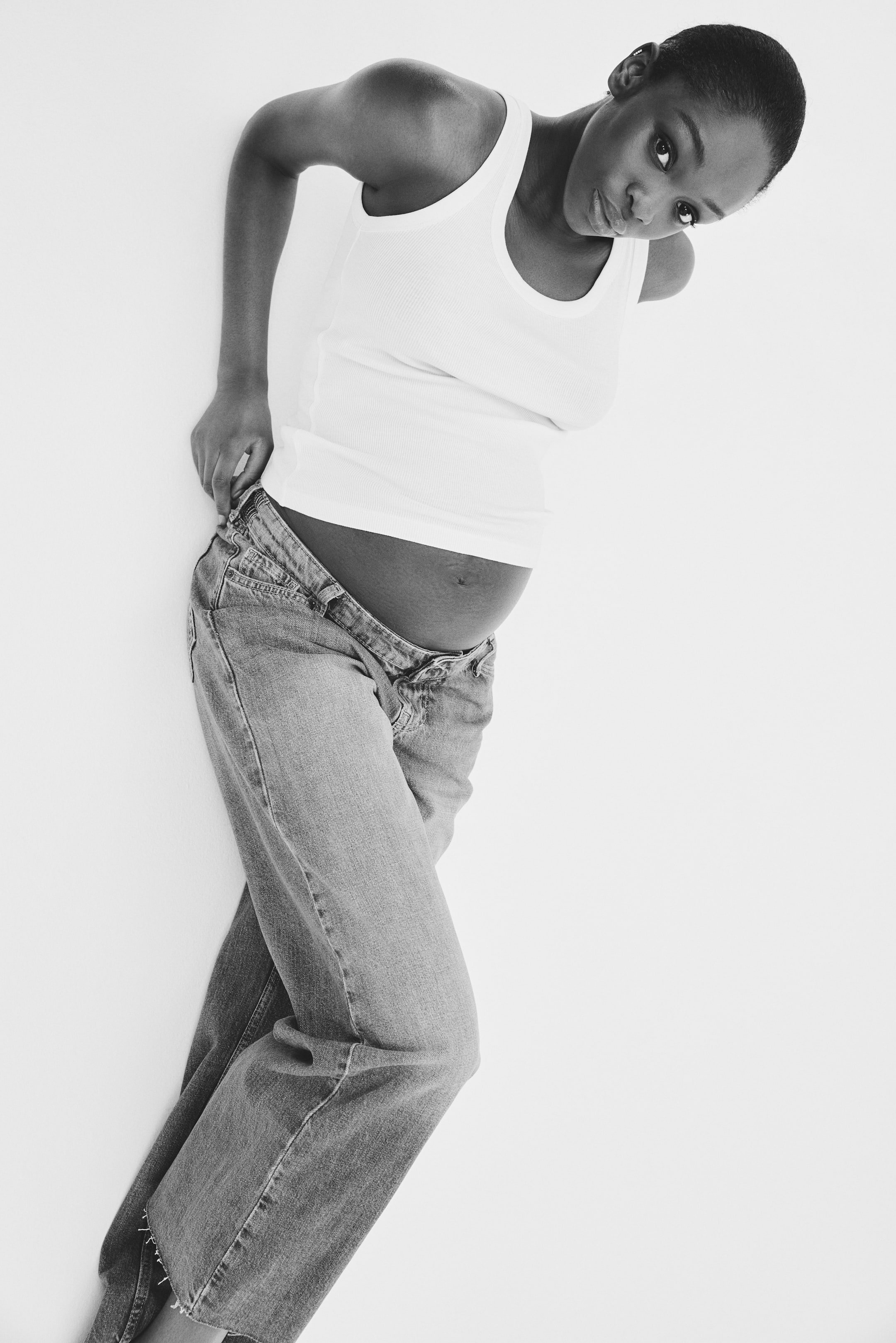 View larger image: A white ribbed vest top is being worn, featuring a scoop neck and a close-fitting silhouette. Light-wash denim jeans are also being worn, with the waistband unbuttoned and unzipped, sitting low on the hips.