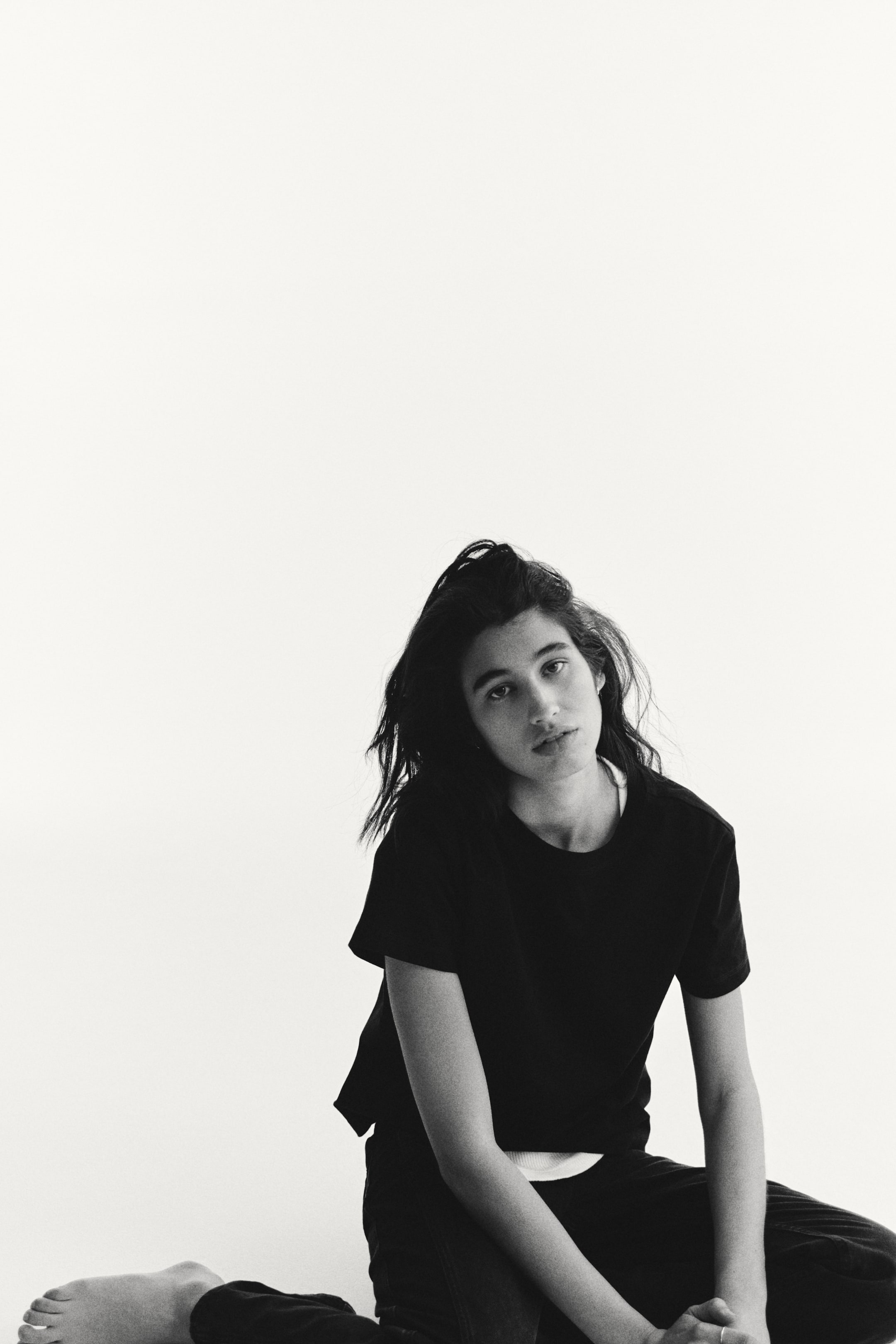 View larger image: A black crew neck t-shirt is worn from a front-left angle, showcasing short sleeves and a relaxed fit. A white undershirt peeks out beneath its neckline and hem, while black relaxed-fit trousers are also worn.