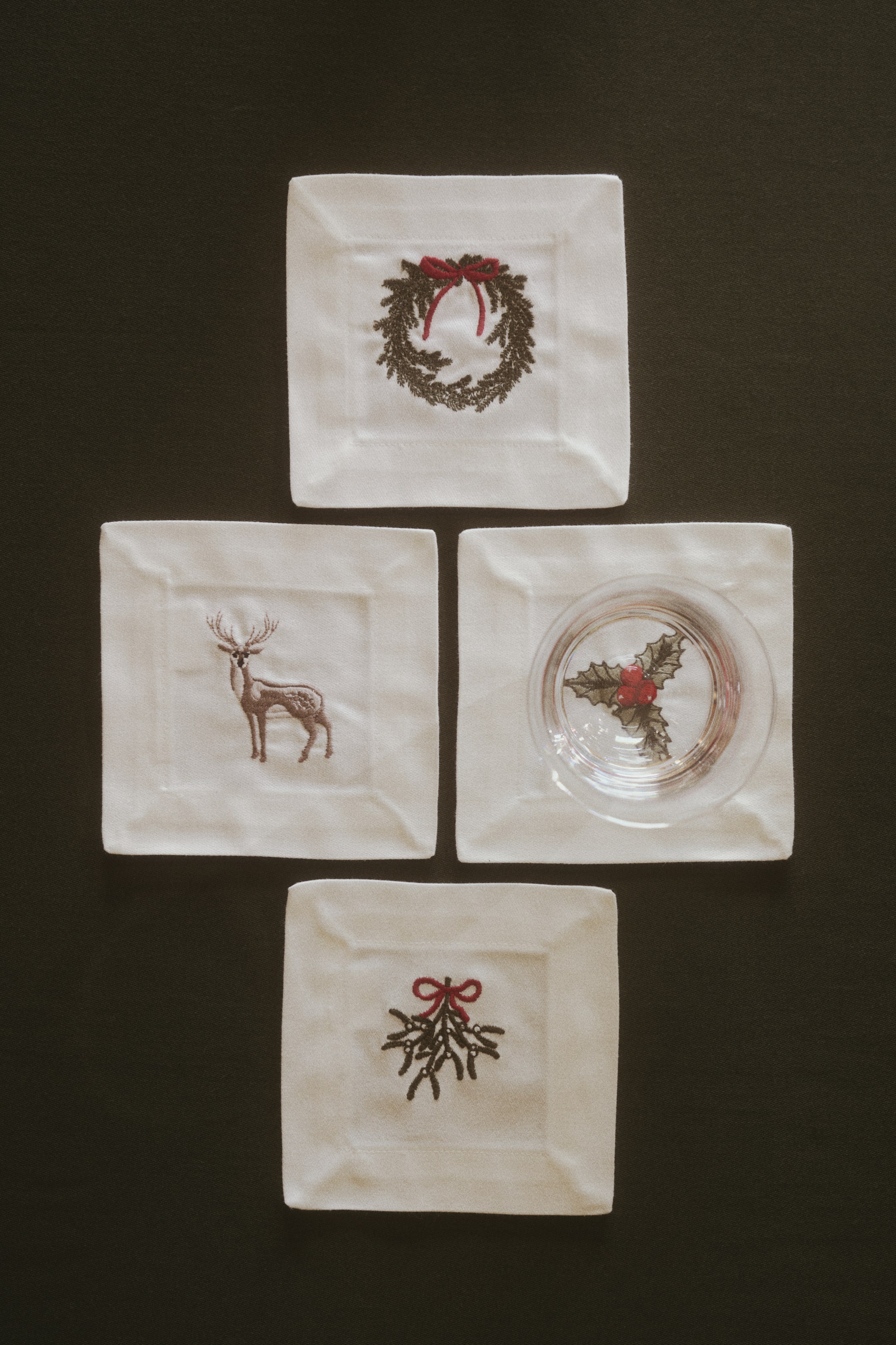 Agrandir l'image: Quatre sous-verres carrés en coton sateen blanc cassé mettent en valeur différents motifs festifs brodés, notamment une couronne, un cerf, du gui et un rameau de houx. Chaque sous-verre présente un large ourlet replié, avec un verre tumbler transparent posé sur le sous-verre de droite.