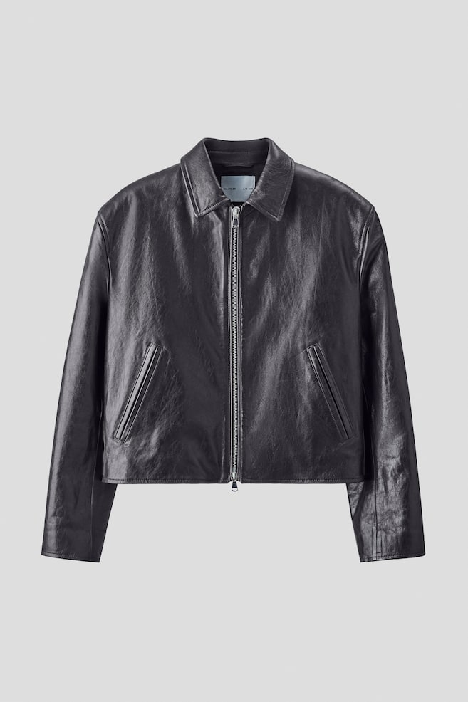 A dark gray leather jacket is presenting its FRONT, featuring a short, boxy, loose-fitting style with a natural grain texture, a collar, a two-way zip down the front, long sleeves, and diagonal jetted front pockets. - 1