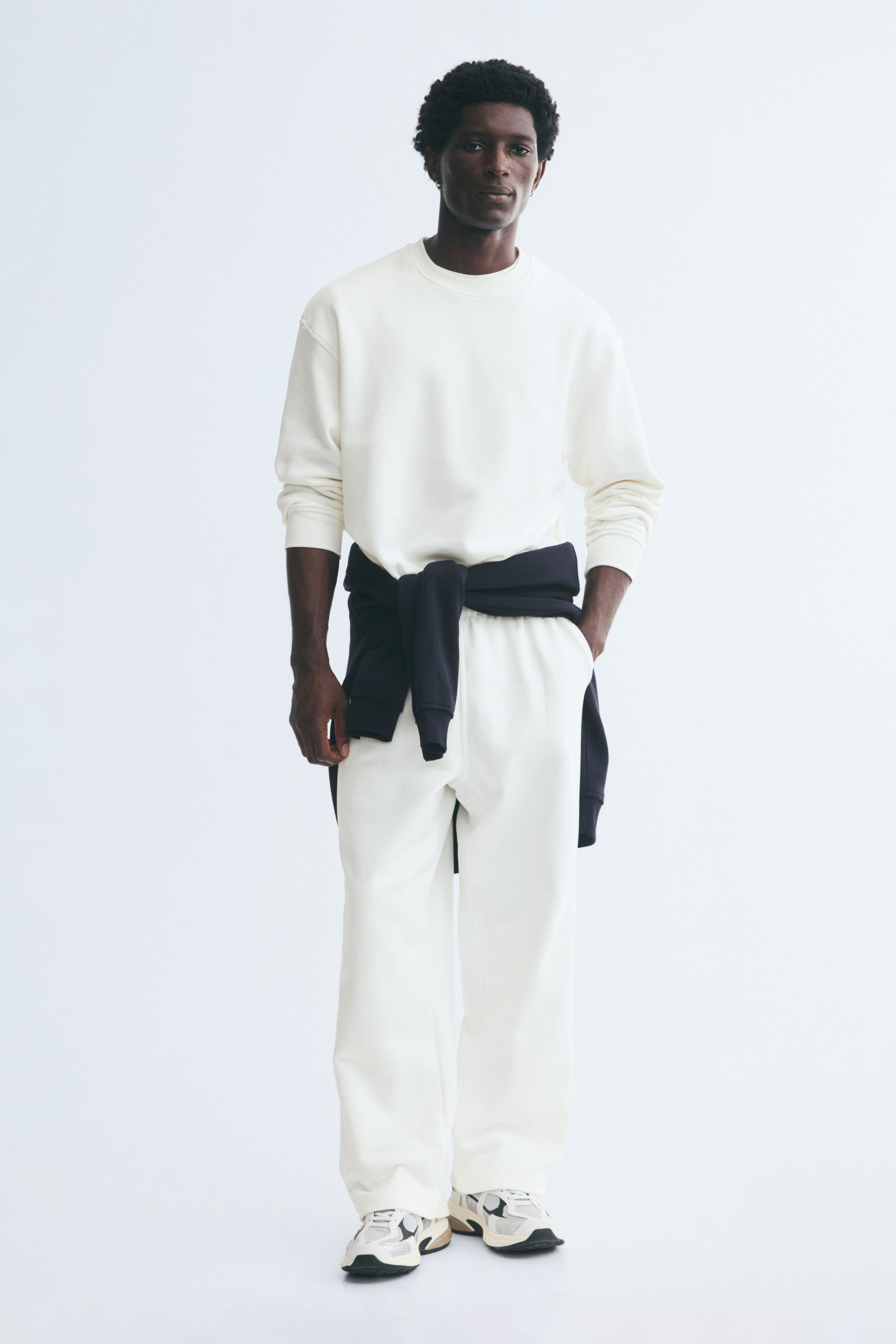 Xem ảnh lớn hơn: Quần track pants bằng vải nỉ cotton màu trắng ngà được người mẫu mặc, với phom rộng thoải mái và túi bên. Người mẫu đang đứng với một tay đặt trong túi bên, phối quần cùng áo sweatshirt cổ tròn đồng bộ và một chiếc áo len tối màu buộc quanh eo.
