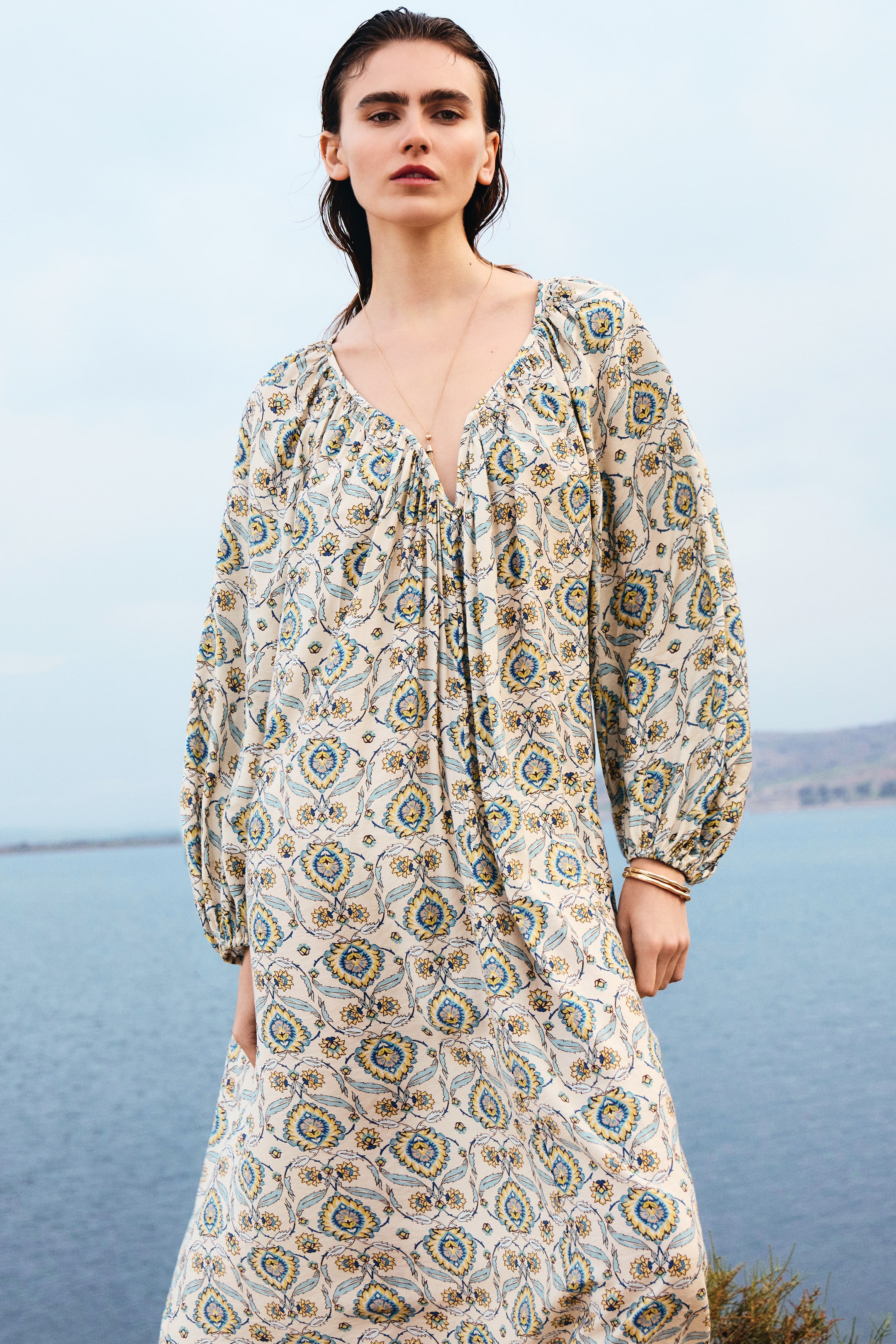 View larger image: The model is showcasing a long, oversized tunic dress crafted from a woven viscose blend fabric, adorned with a vibrant blue and yellow floral pattern. This dress boasts a sweetheart neckline accentuated with binding and gathers, complemented by raglan balloon sleeves with elasticated cuffs. Additionally, the model is accessorized with a gold-toned necklace and bracelet, adding a touch of elegance to the ensemble.