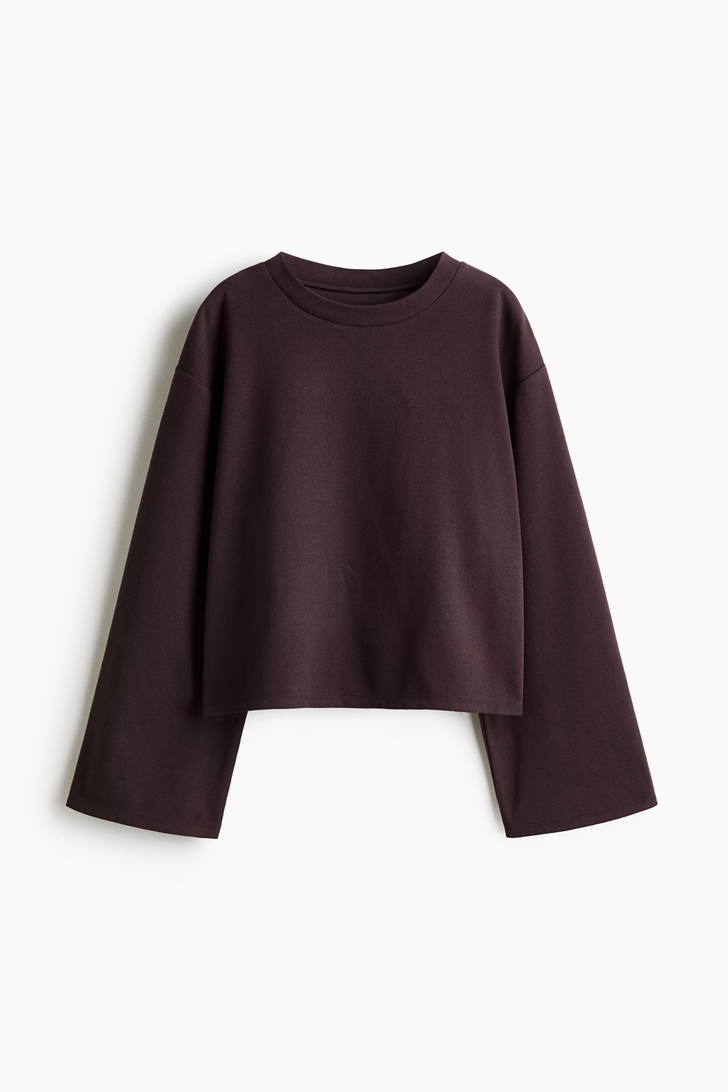 A dark plum jumper in brushed fleece fabric is being worn by the model, featuring a round neck, dropped shoulders, and long, wide sleeves. The jumper is layered over a white T-shirt and paired with beige trousers as the model is leaning forward.