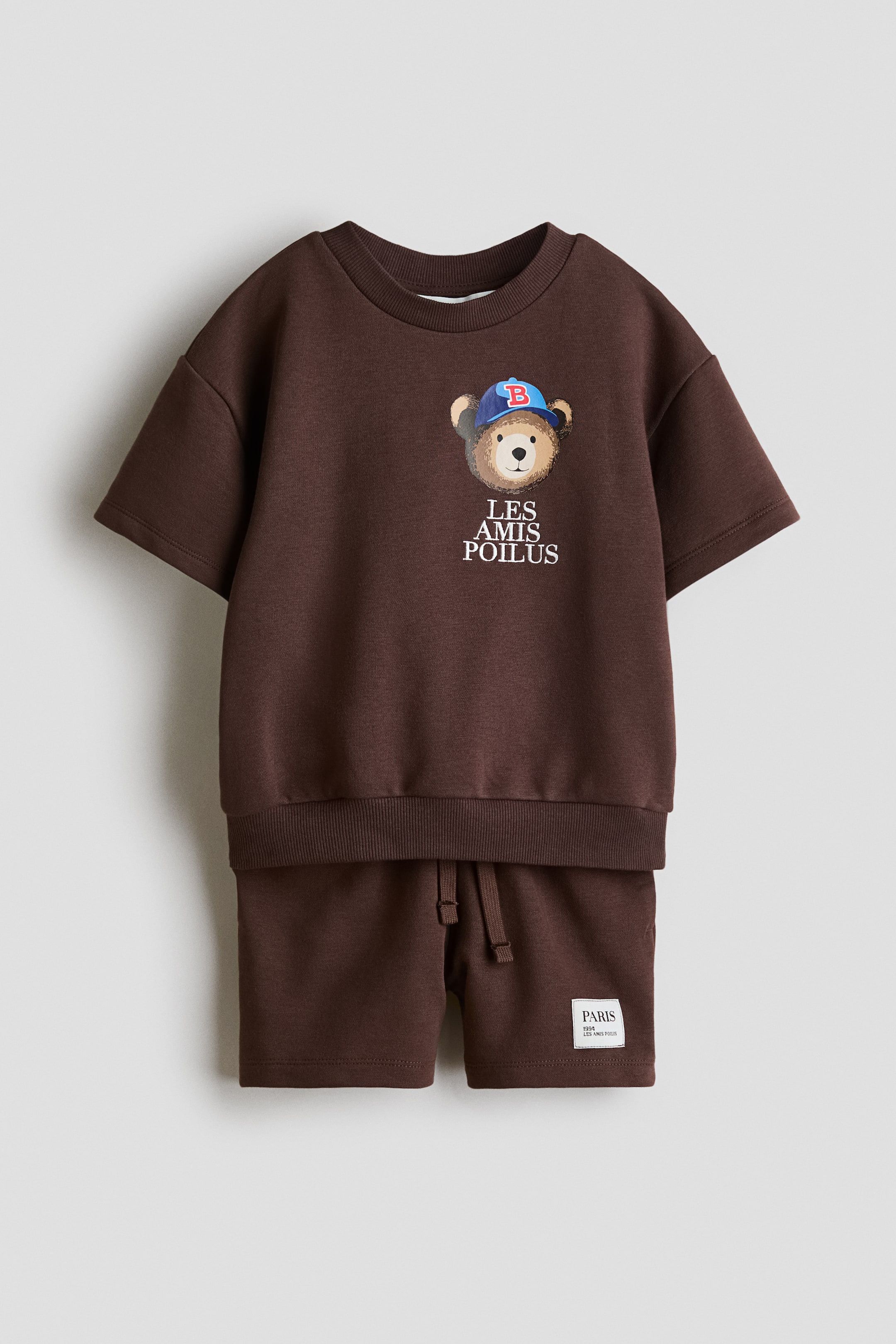 View larger image: A brown two-piece set showcases a short-sleeved top and matching shorts from the front. The top features a round, rib-trimmed neckline, dropped shoulders, ribbing at the hem, and a chest graphic of a bear wearing a blue cap above the text "LES AMIS POILUS". The shorts have an elasticated, drawstring waist and a small white label on the left leg.