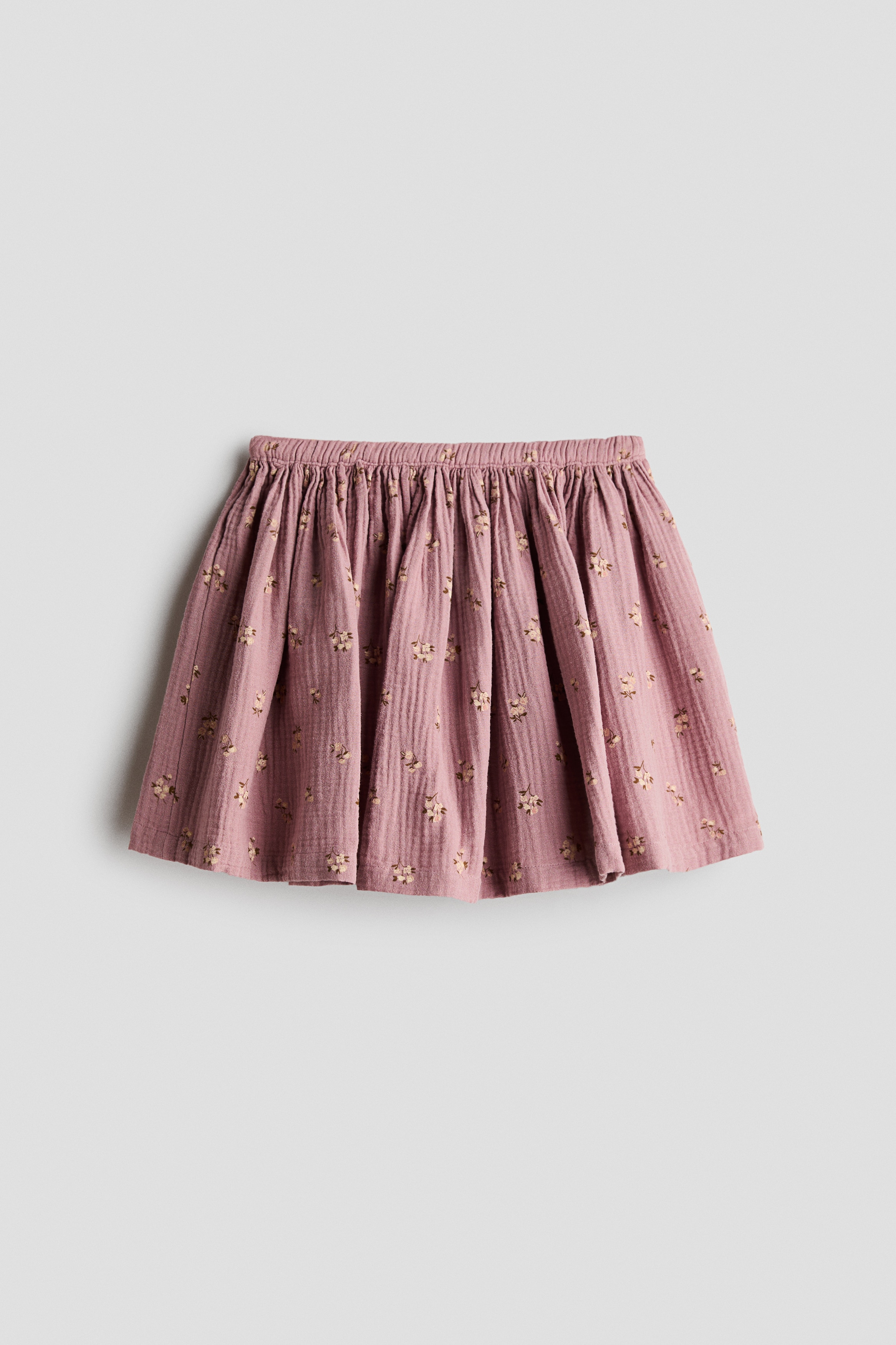 A flared skirt in dusty pink cotton muslin with an all-over floral print is displayed from the front. The skirt features a gathered waist that adds volume and is styled with a white ribbed T-shirt.