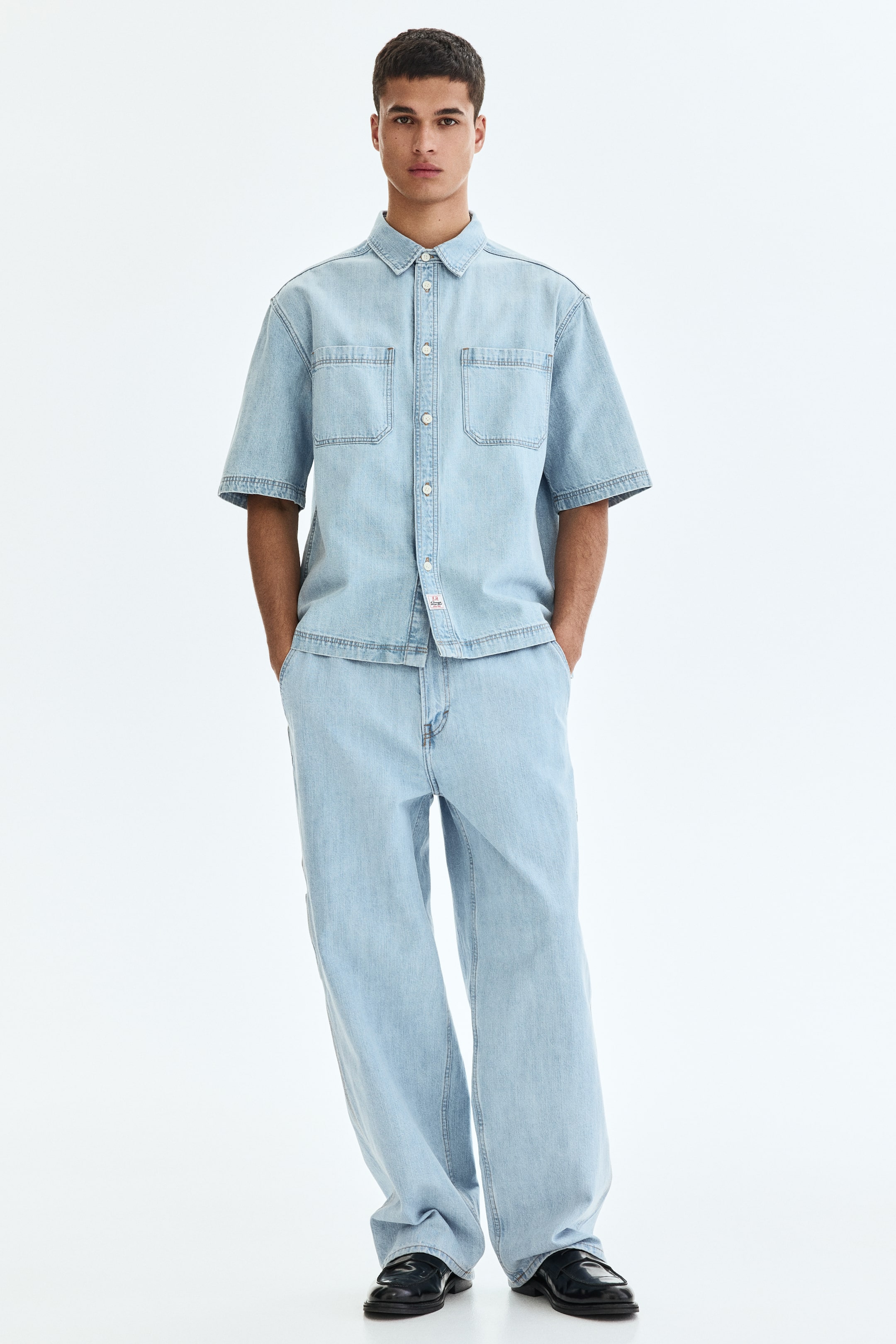 Vezi imaginea mai mare: O cămașă din denim cu mâneci scurte și spălare ușoară, având un guler clasic, o închidere completă cu nasturi albi și două buzunare aplicate pe piept, toate accentuate cu cusături contrastante vizibile. Pantalonii asortați, din denim cu spălare ușoară și croială largă, prezintă, de asemenea, cusături contrastante, având o croială relaxată care se așază peste pantofi de tip loafers negri.