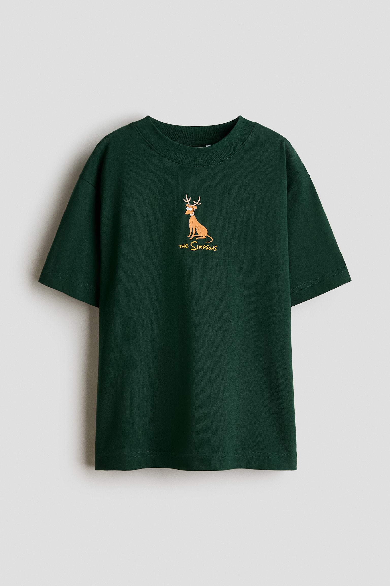 A dark green cotton jersey T-shirt is shown hanging in a front-facing view, featuring a round rib-trimmed neckline and dropped shoulders. The T-shirt displays a small printed graphic at the centre chest of a cartoon dog wearing antlers above the text "the Simpsons".