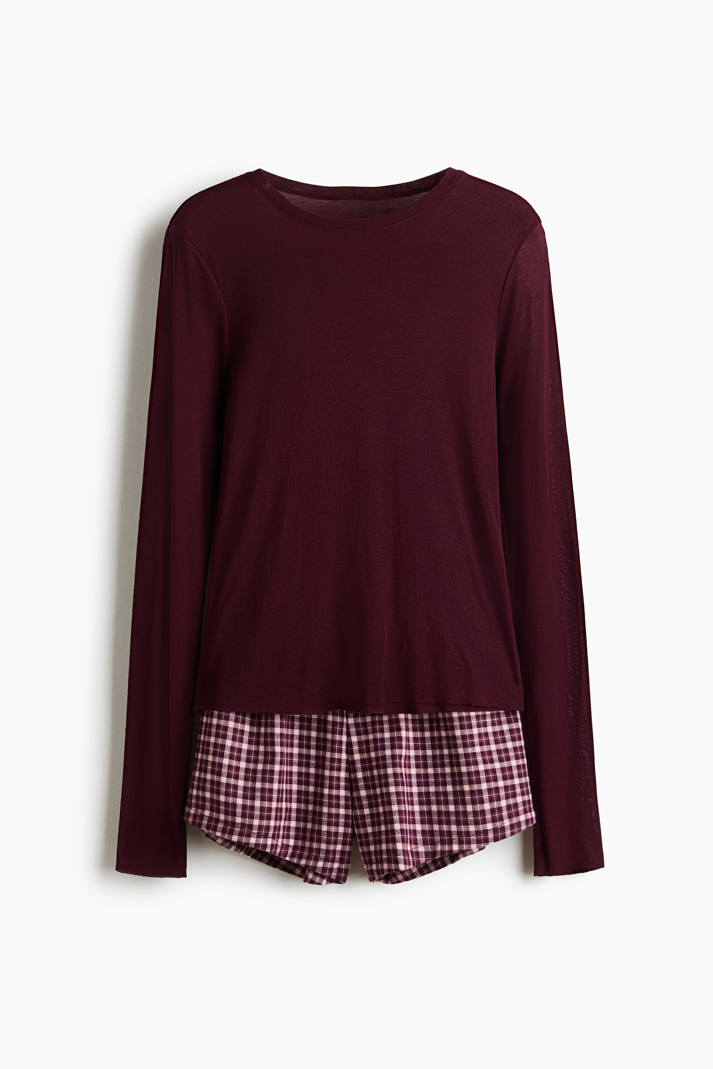 Et pyjamassæt bestående af en langærmet jersey-top i bordeaux og matchende bordeaux- og hvidternede shorts bæres af modellen. Modellen står med armene korslagt over талjen og fremhæver shortsene med elastisk linning og dekorative gylpknapper.