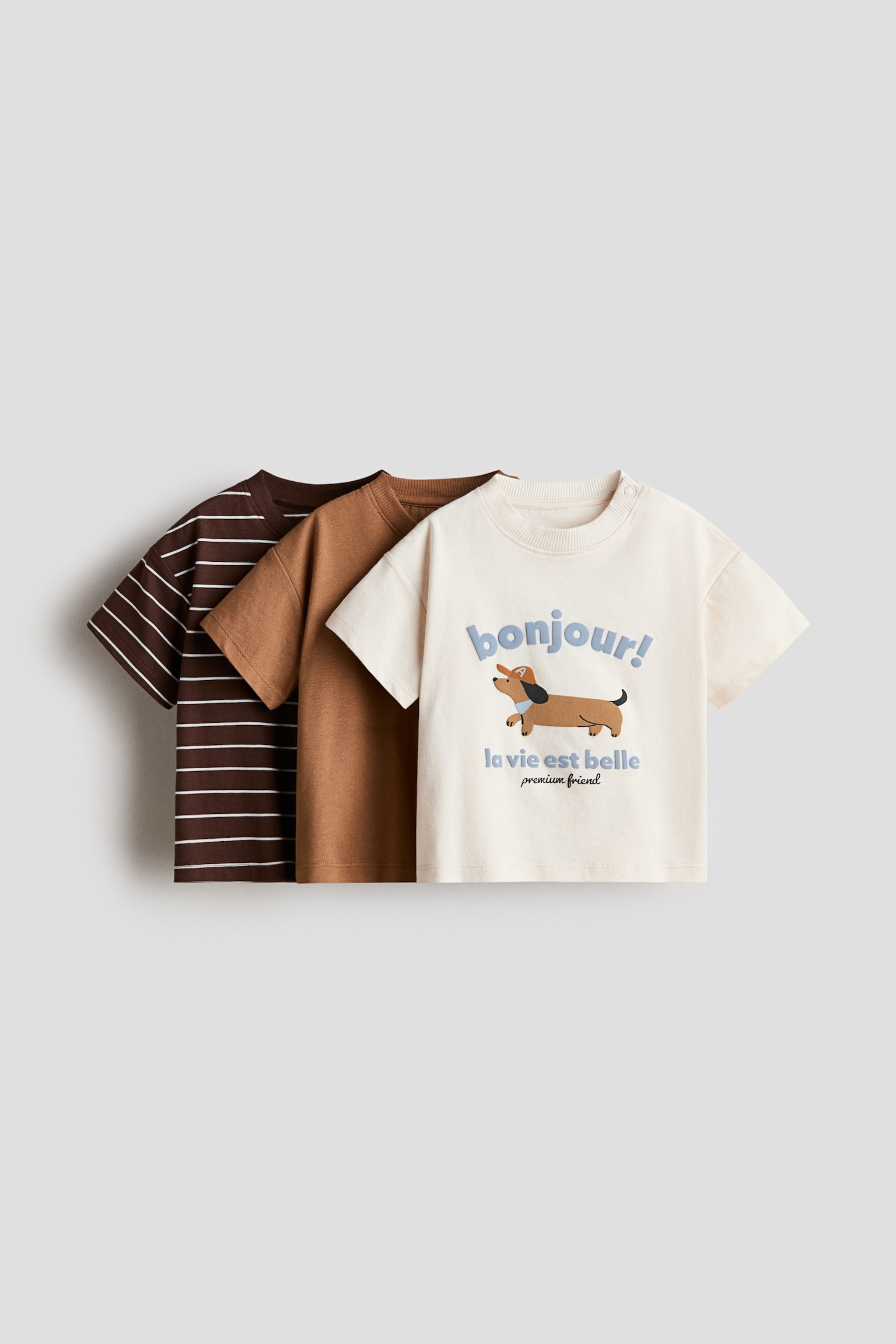 A 3-pack of cotton t-shirts features three short-sleeve crewneck tees layered and facing front. The front cream-coloured tee features a dachshund graphic with the text "bonjour!" and shoulder snap buttons, while the middle tee showcases a solid light brown colour, and the back tee displays a dark brown and white striped pattern.
