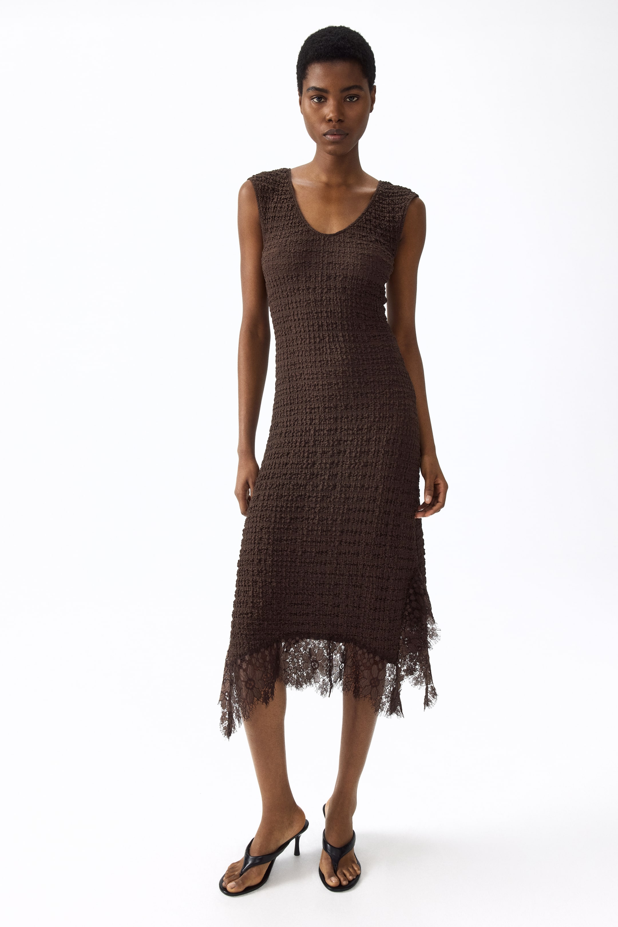 Ver imagem maior: Um vestido midi castanho sem mangas apresenta um tecido de malha texturizada, um decote em V e uma silhueta justa. Apresenta uma bainha assimétrica com um acabamento em renda escura e desfiada.