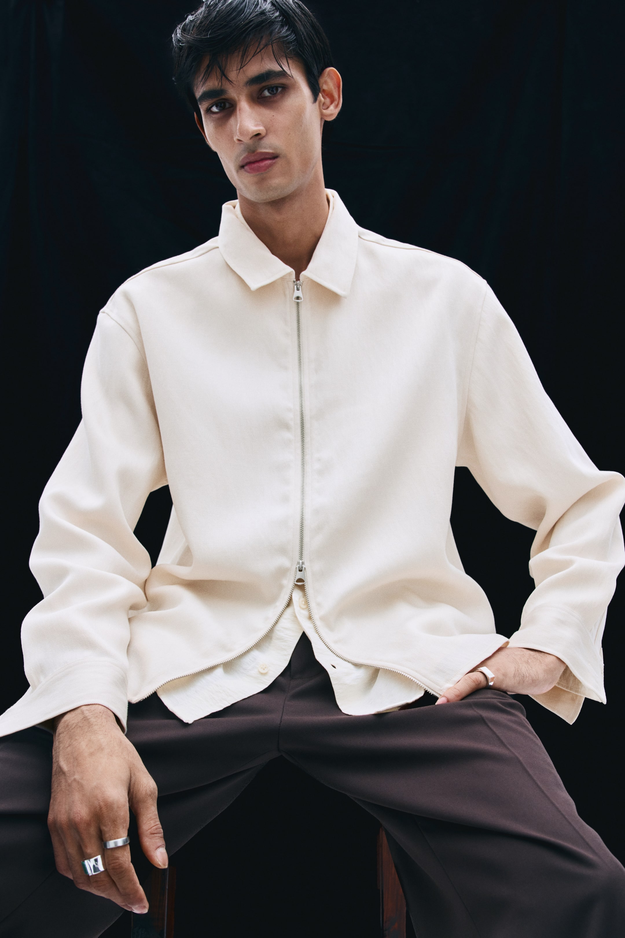 View larger image: The model is wearing an off-white twill overshirt featuring a classic collar, long sleeves, and a silver two-way front zipper. The overshirt is styled partially unzipped at the hem, layered over a light-coloured button-up shirt and paired with dark brown trousers.