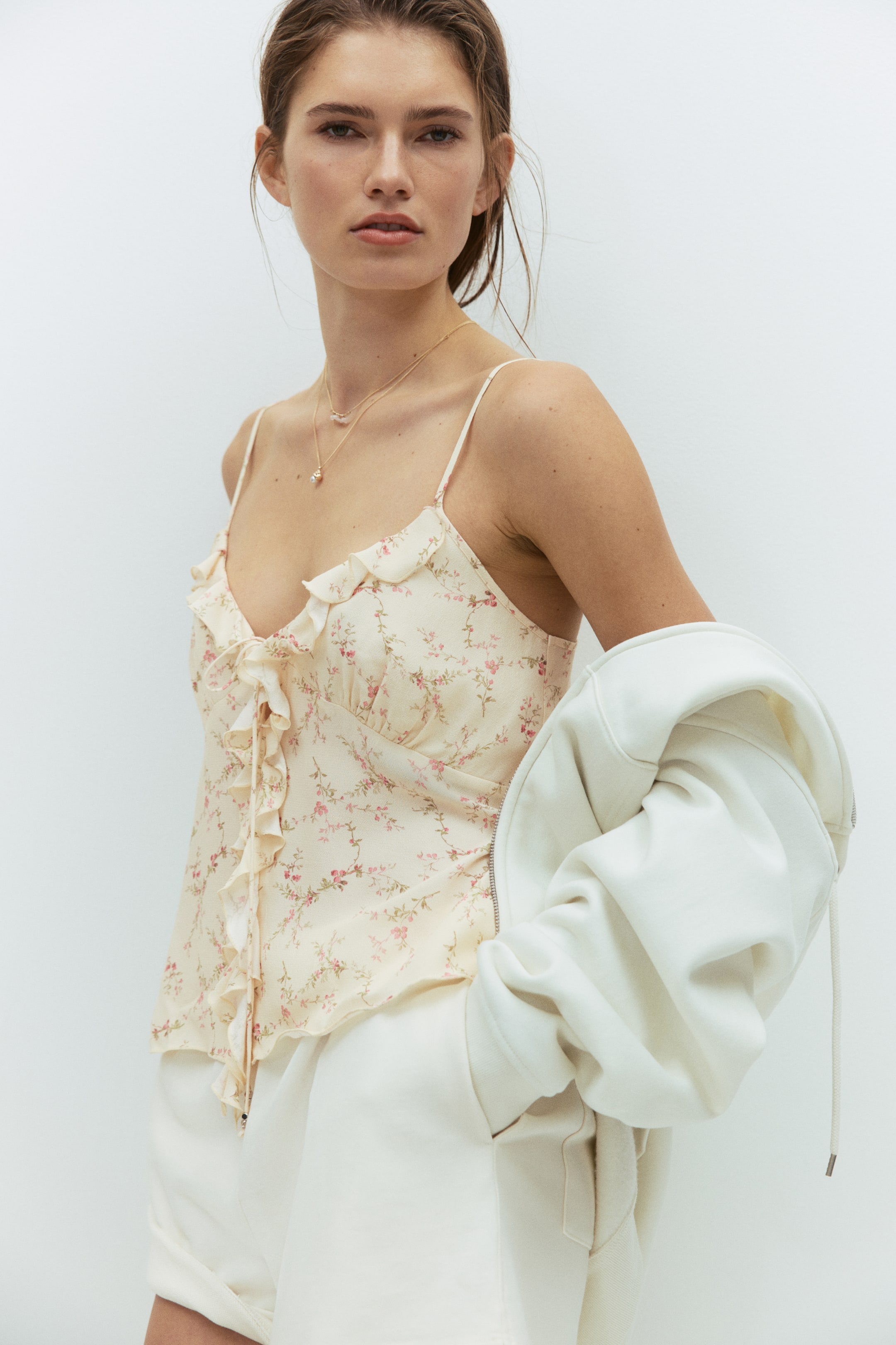 View larger image: A pale yellow floral print viscose top, featuring flounce trim along the V-neckline and down the front with lacing, is worn by the model. A white jacket is draped over one of the model's arms, partially covering the top, and a delicate gold layered necklace is worn over the neckline.