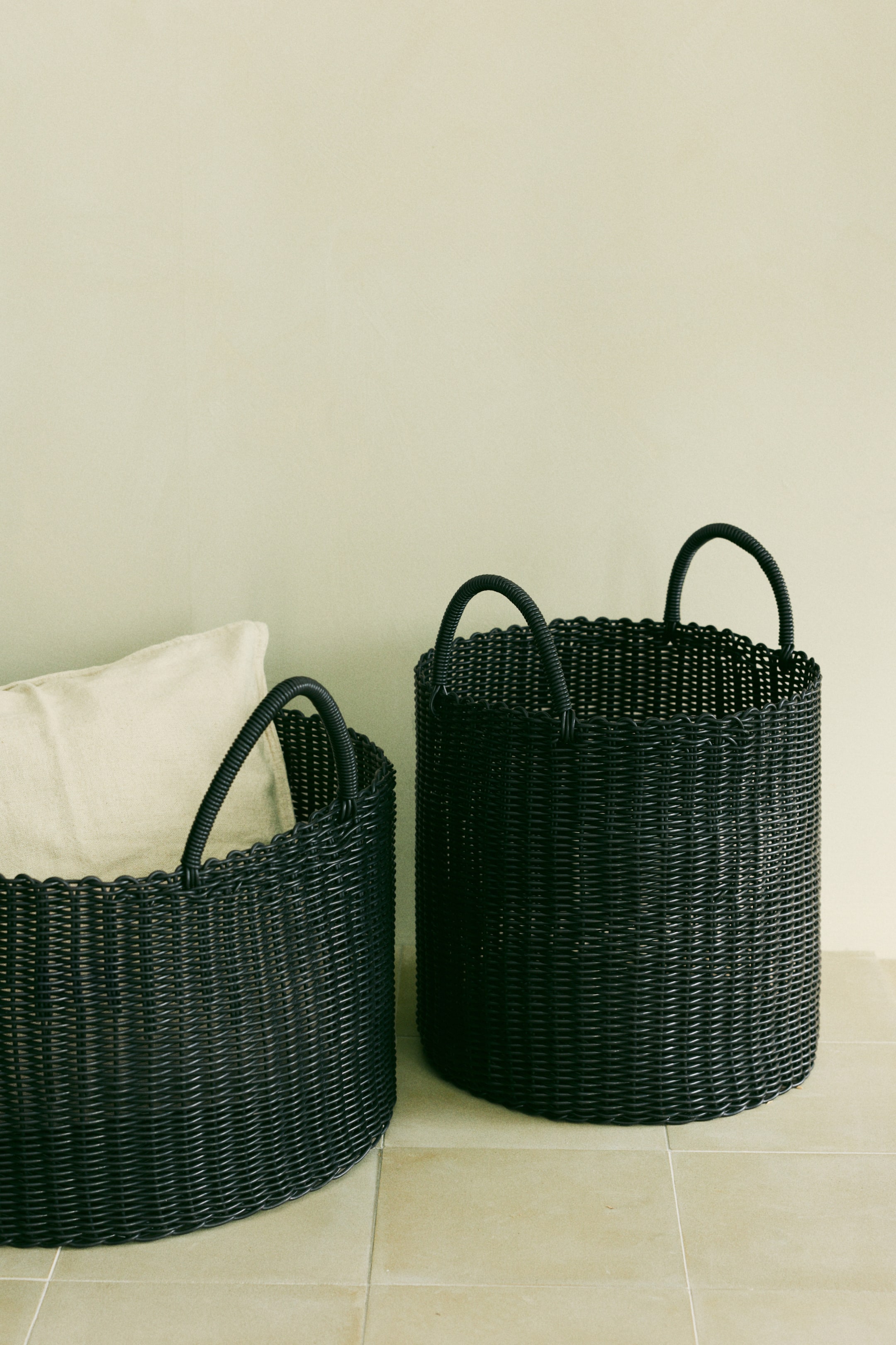 Visa större bild: Två handvävda svarta plastförvaringskorgar, båda med två böjda handtag upptill, står framåtvända; den mindre korgen till vänster håller en ljus kudde, medan den större korgen till höger är tom.