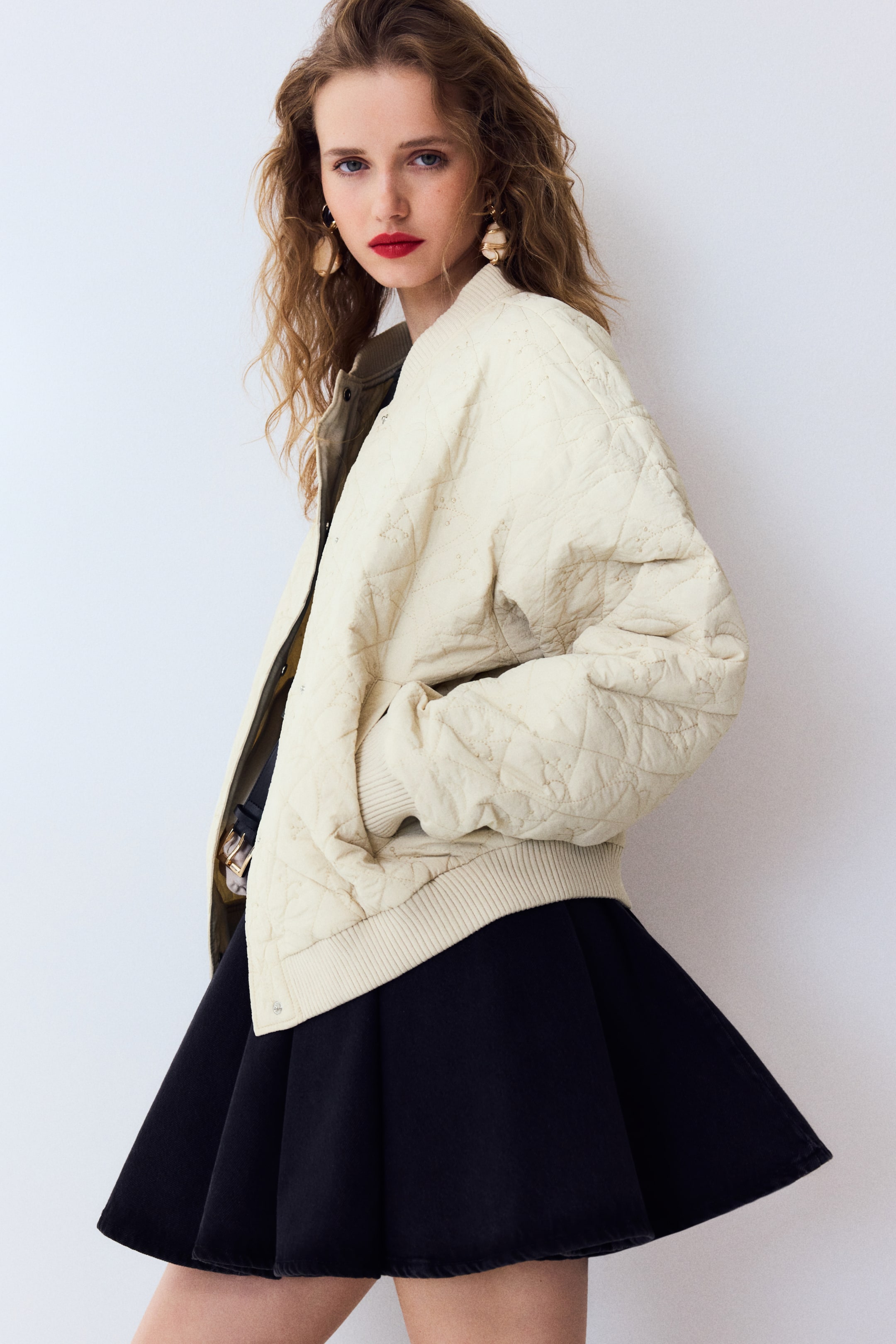 View larger image: A cream quilted bomber jacket ditunjukkan dari sudut hadapan-sisi sedikit, menampilkan corak quilt berlian keseluruhan dengan perincian sulaman kecil, kolar berusuk, manset berusuk, dan hem berusuk yang terletak di atas skirt gelap. Satu tangan diletakkan dalam poket sisi.