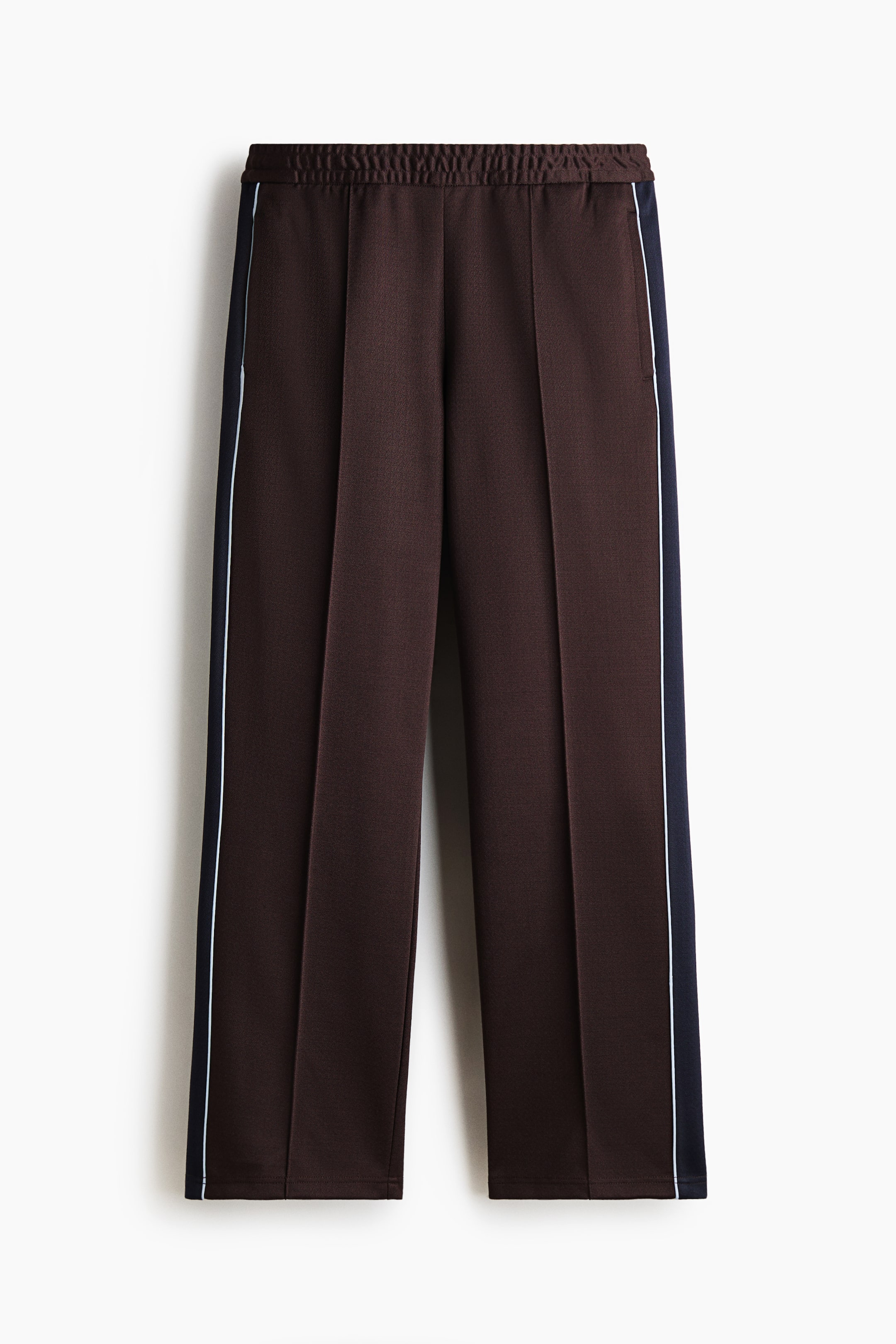 View larger image: The brown trousers are featuring an elasticated waistband and a prominent central front crease on each leg. A dark blue side stripe with a thin white line is running vertically along the outer seam of each leg.