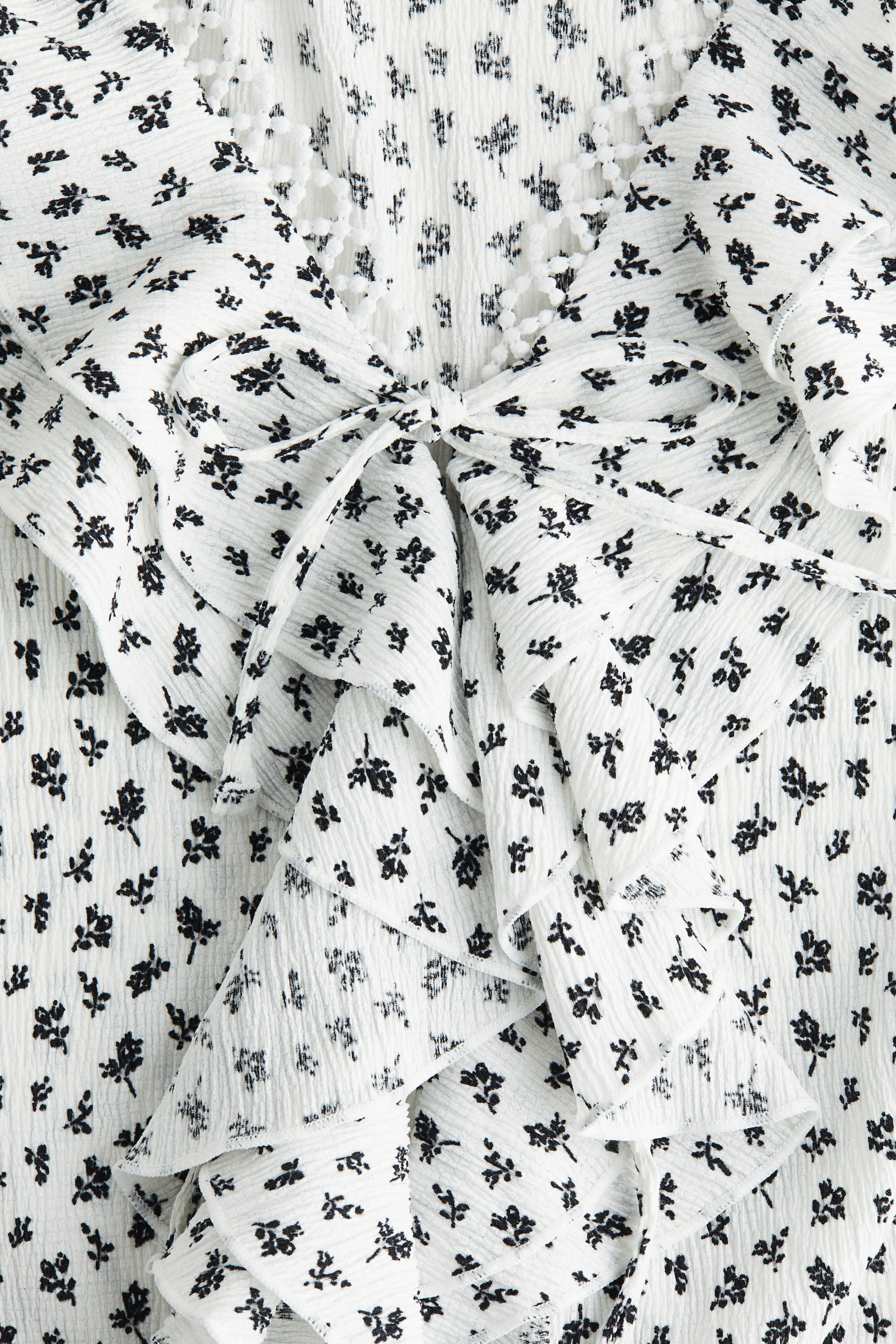 View larger image: A white, crinkled top with a black floral print is featuring a tie-front closure, with cascading ruffles adorning the front opening. A white bobble trim is detailing the V-neckline.