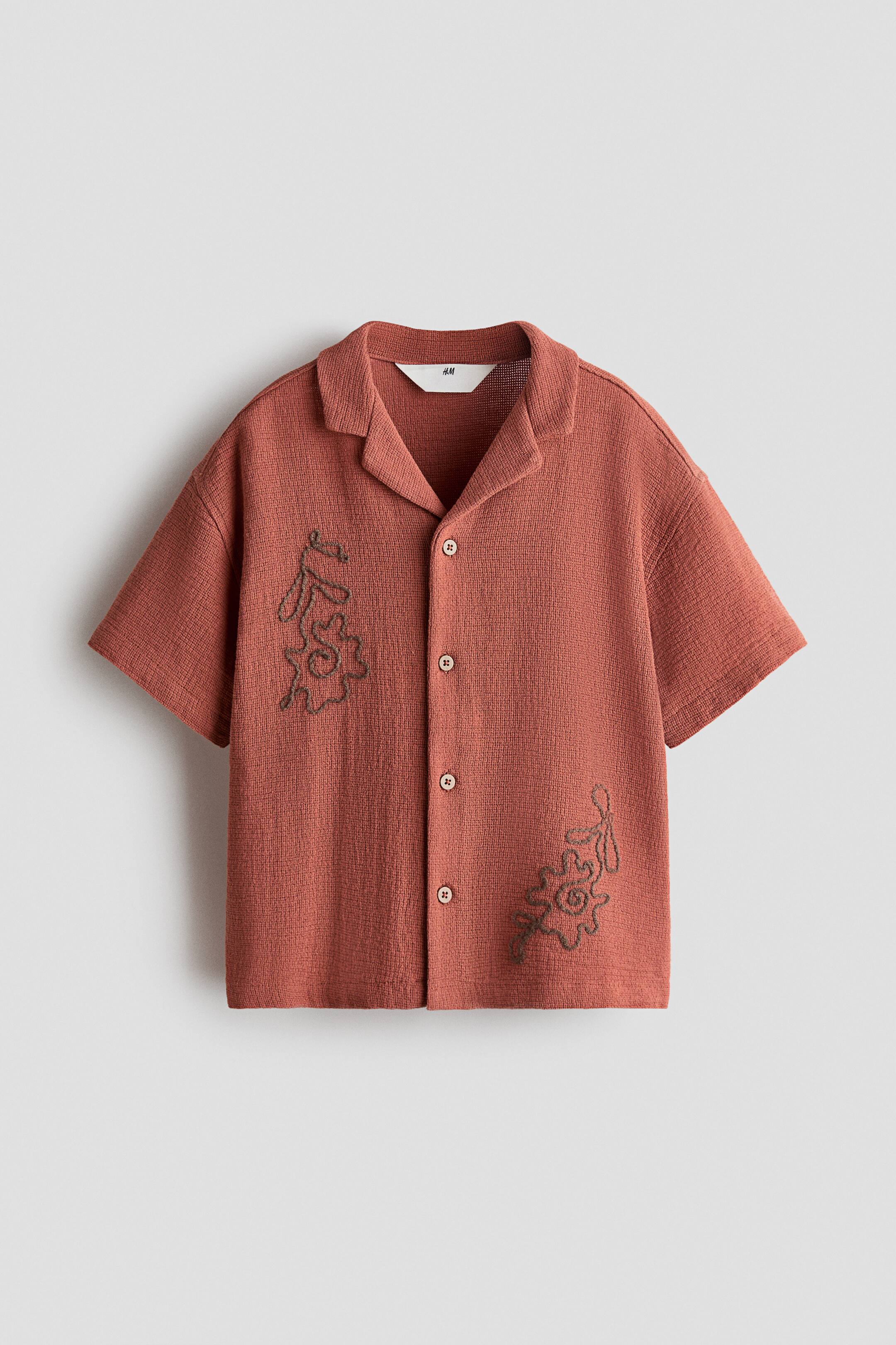 View larger image: A terracotta-coloured short-sleeve shirt in a textured cotton weave is shown from the front, featuring a resort collar, dropped shoulders, and a button-down closure. The shirt displays dark grey embroidered abstract floral motifs on the upper right chest and lower left front.