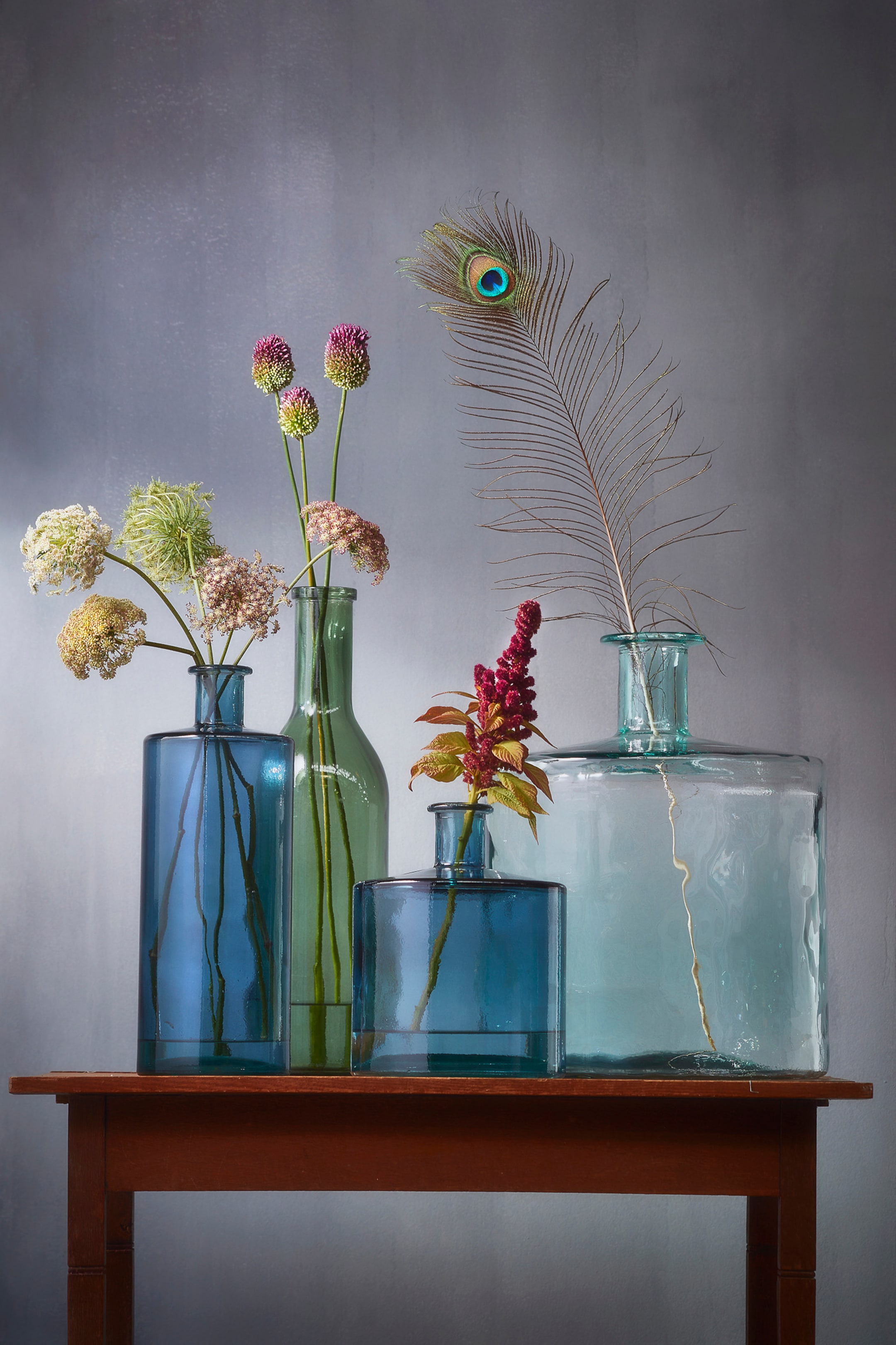 Se større billede: En samling af fem glasbeholdere, der varierer i form, størrelse og farve, viser forskellige botaniske arrangementer. En høj, rektangulær blå flaske holder mørke stilke, mens en kortere klassisk blå flaske bagved præsenterer hvide og grønne blomster. En klassisk grøn flaske fremviser lilla-lyserøde allium-lignende blomster, og en kort rektangulær blå flaske indeholder rød amarant. En bred, firkantet lyseblå flaske med en kort hals fremviser en påfuglefjer.