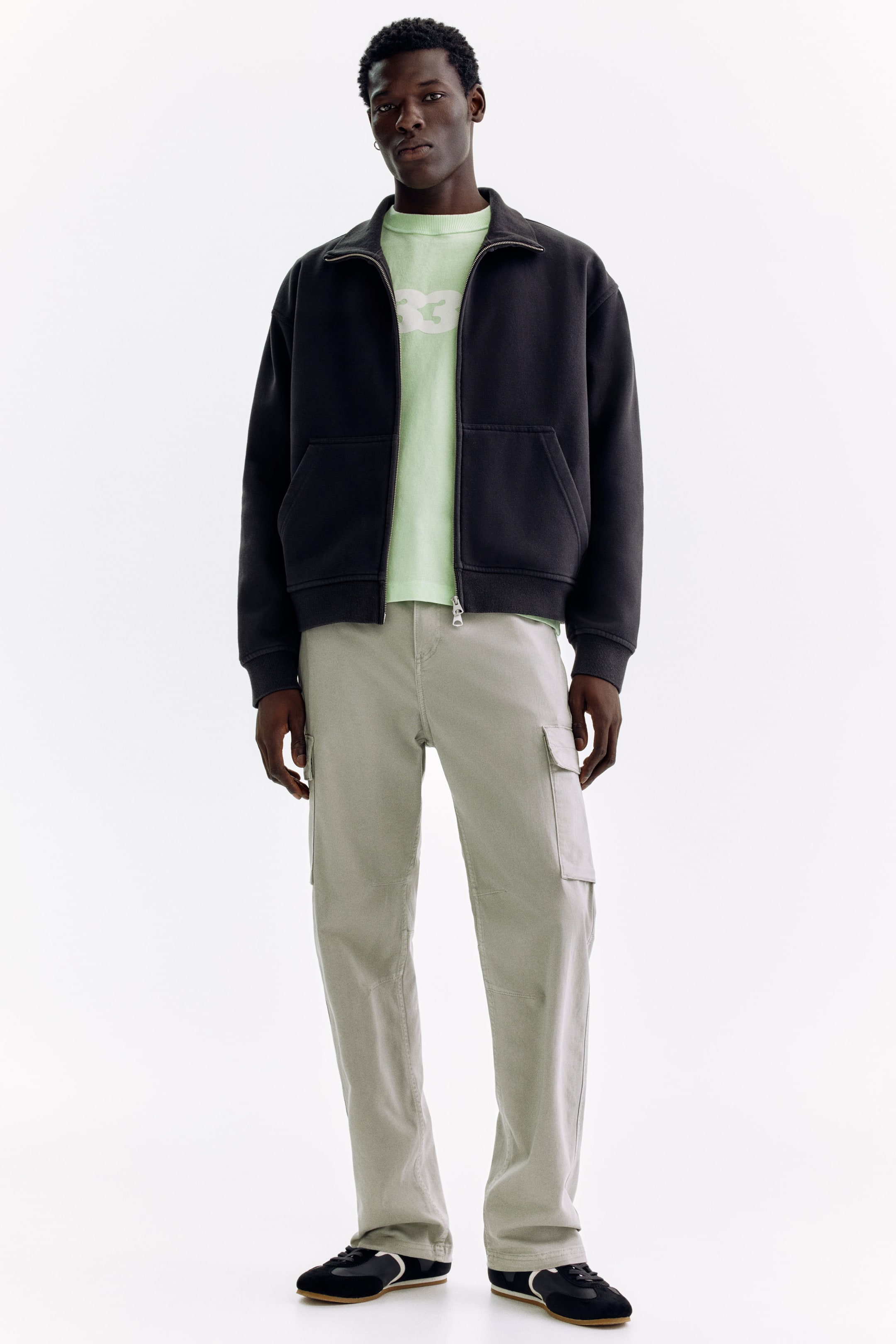 View larger image: Presenting a complete ensemble, showcasing a dark charcoal full-zip jacket with a stand-up collar and front pockets, layered over a pale green graphic t-shirt. The look is completed with light sage green cargo pants featuring side pockets, paired with black sneakers accented by white trim.