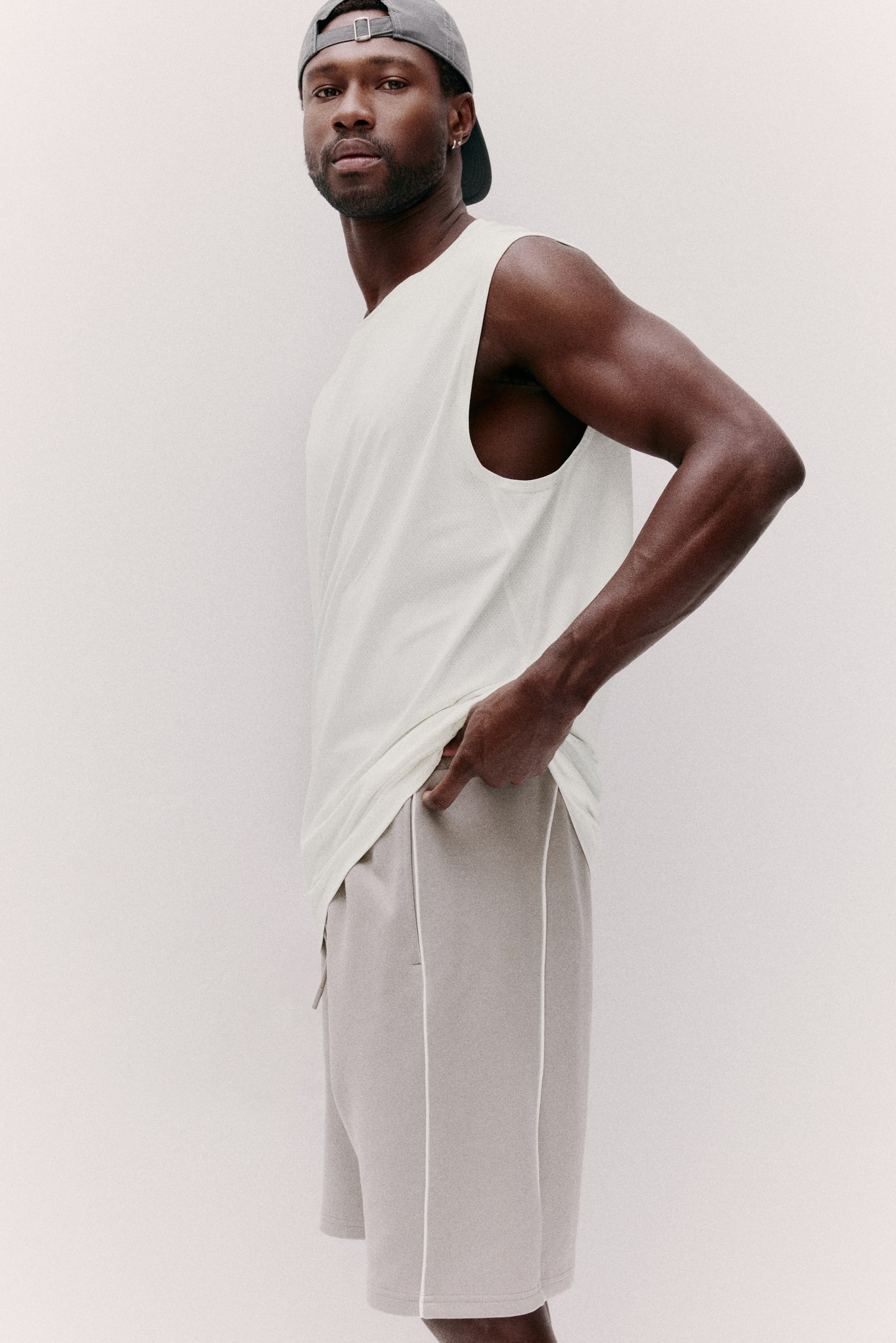 View larger image: A cream-colored sleeveless tank top, featuring a relaxed fit and round neckline, is paired with light gray knee-length shorts detailed with white vertical piping along the sides and a drawstring at the waist. A gray baseball cap is worn backward, showcasing its metal buckle adjuster.