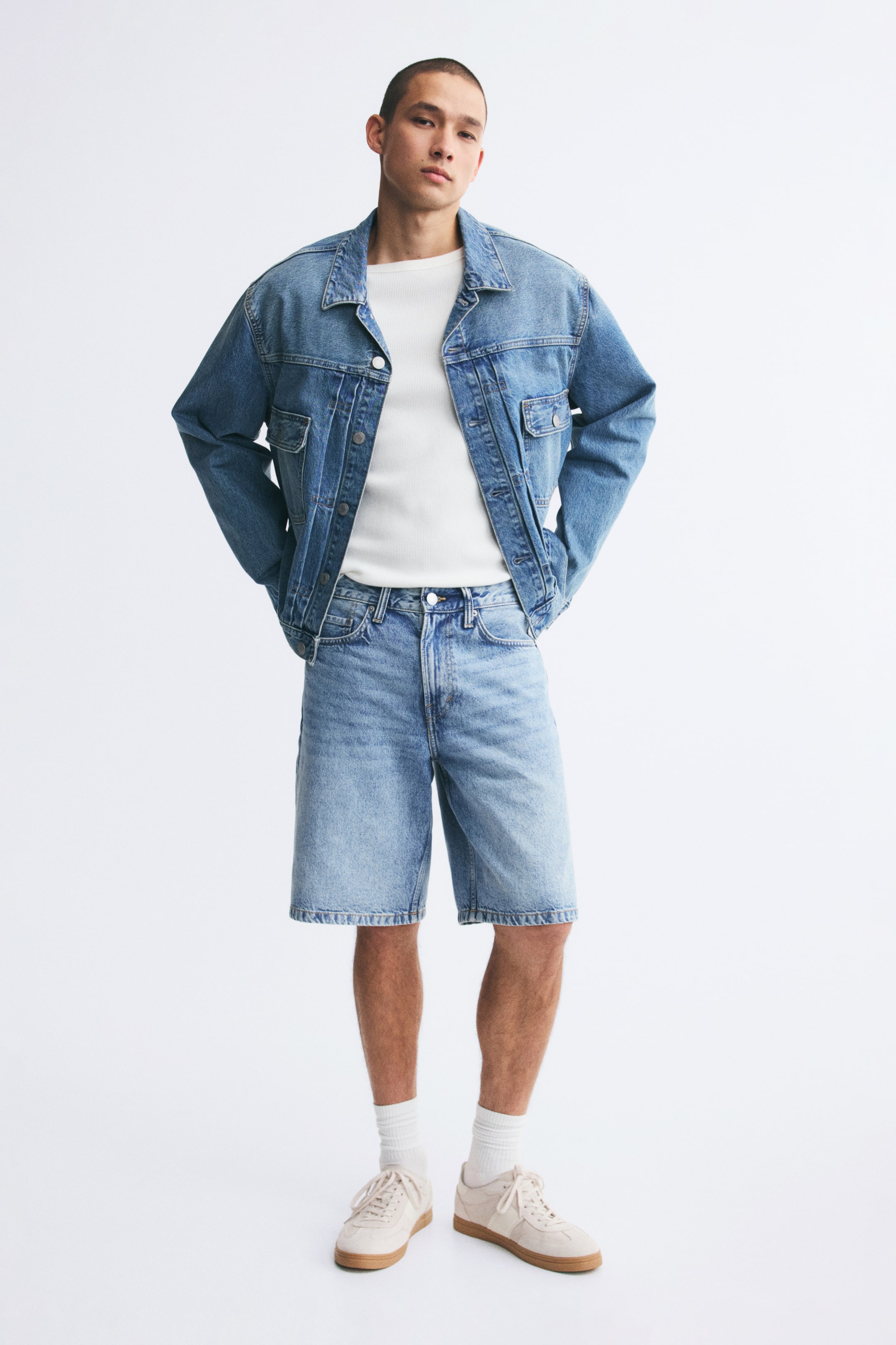 Vezi imaginea mai mare: Pantaloni scurți din denim cu spălare deschisă, cu croială lejeră și stil cu cinci buzunare, purtați de model. Pantalonii scurți, care se opresc puțin sub genunchi, sunt asortați cu o jachetă din denim în ton, un tricou alb și sneakers bej.