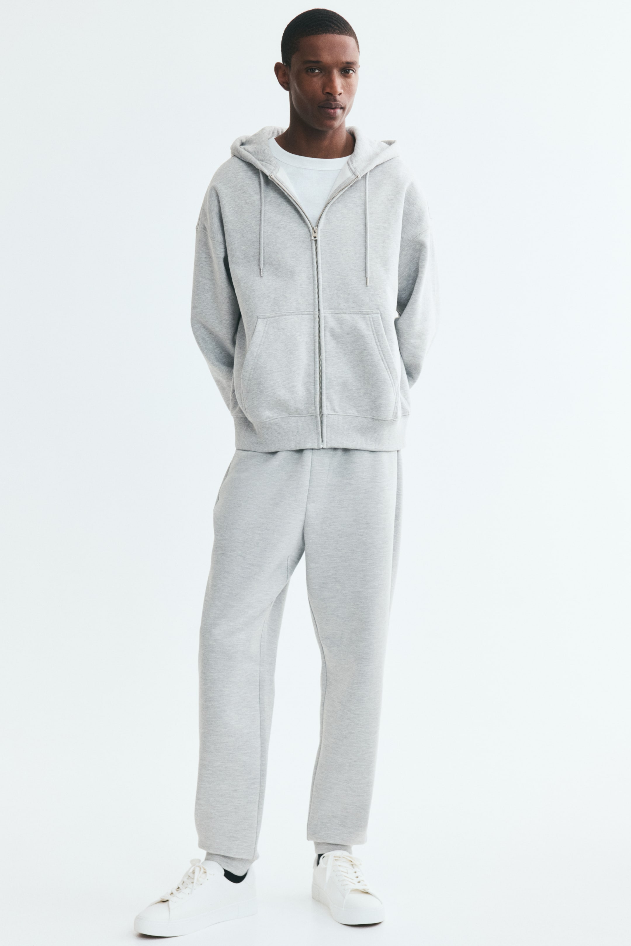 Ver imagen más grande: El modelo lleva puestos unos pantalones de chándal de color gris claro, de corte regular y pernera ajustada, con bajos acanalados. Los pantalones cuentan con bolsillos laterales en diagonal y una bragueta simulada, y se combinan con una sudadera con capucha y cremallera a juego y unas zapatillas blancas.