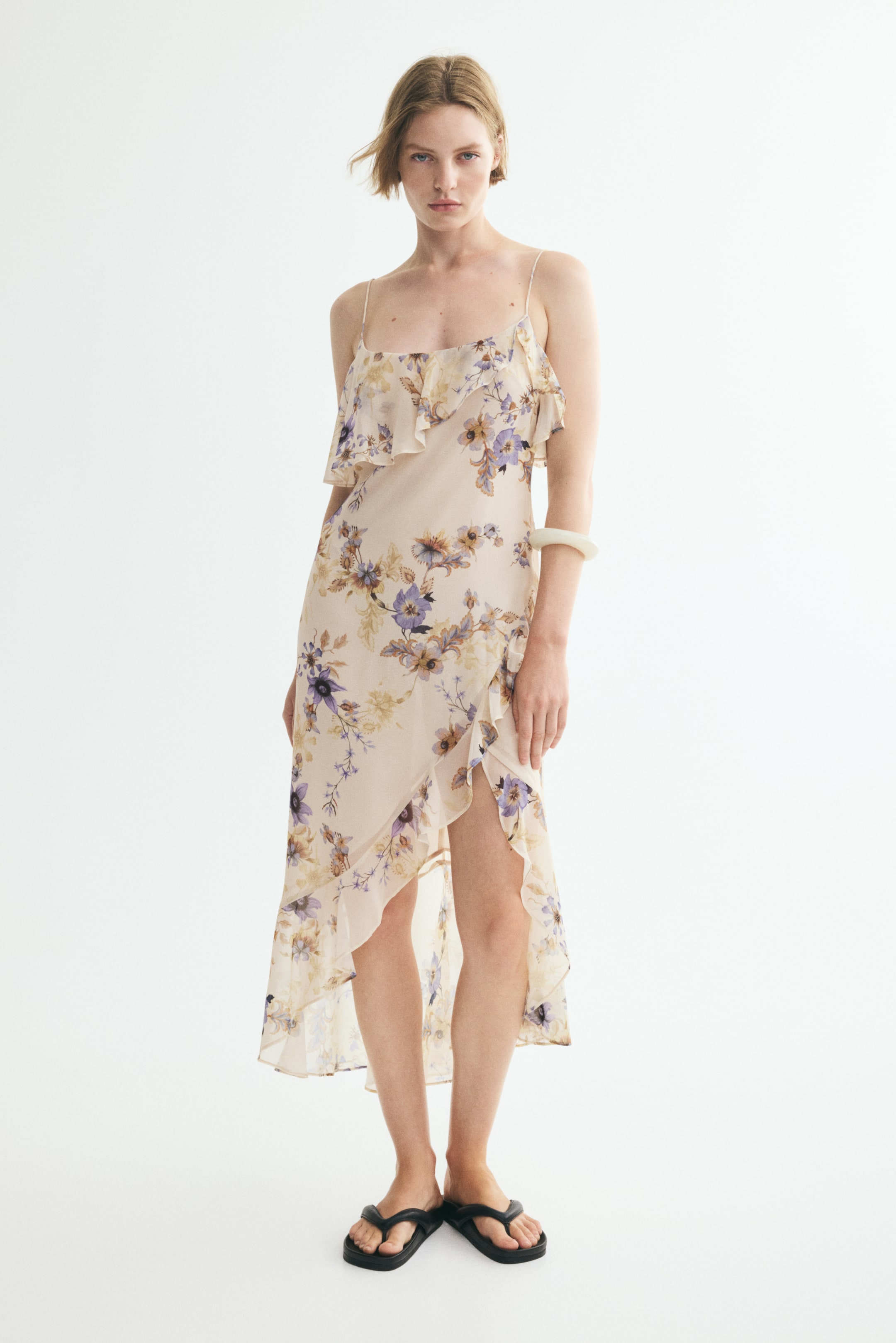 Ver imagem maior: A modelo está a usar um vestido justo de comprimento midi com um padrão floral leve. O vestido apresenta um decote redondo com um folho assimétrico, alças finas ajustáveis e uma saia cruzada com um folho largo na bainha.