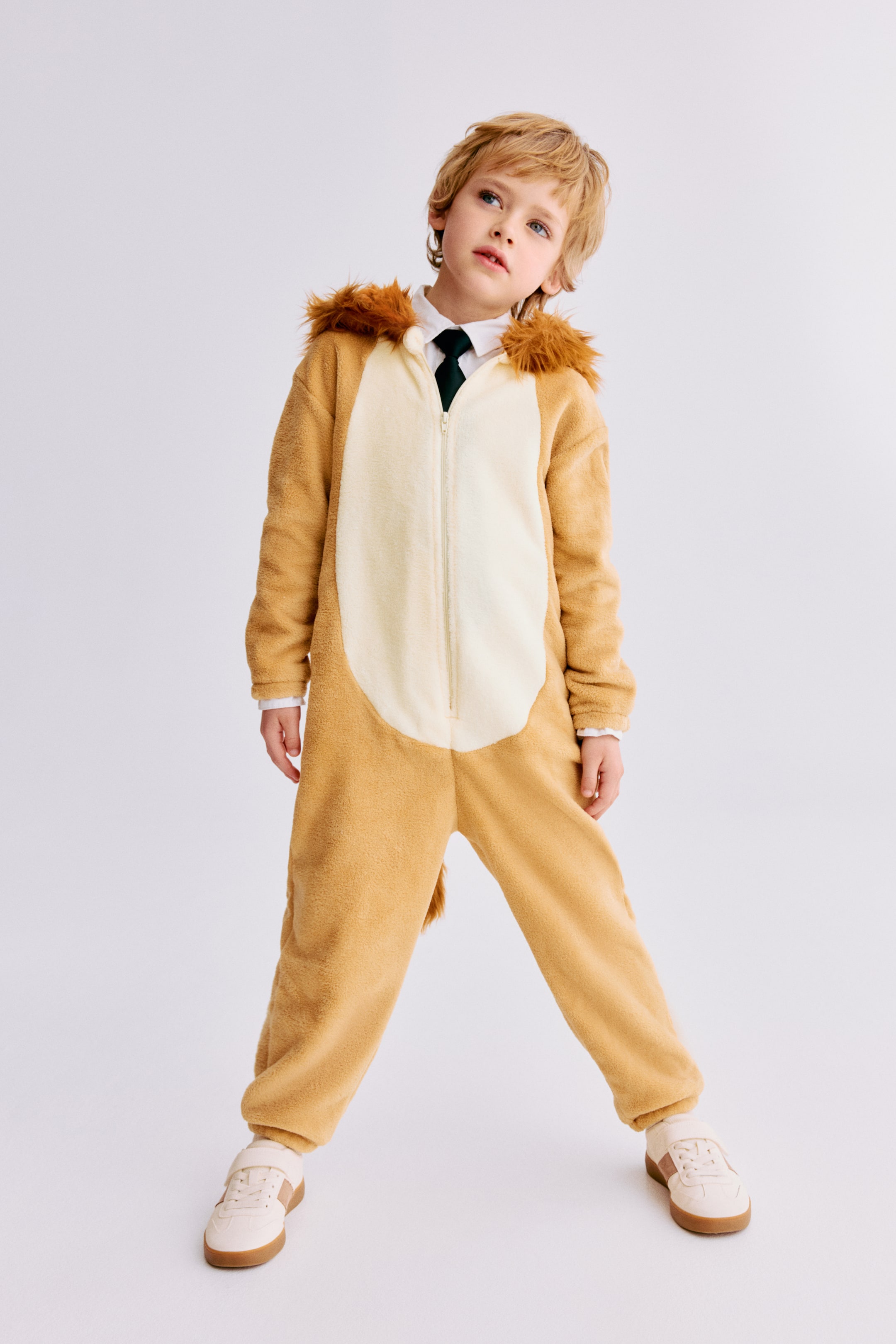 Ampliar la imagen: Un traje de cuerpo completo con temática de león se presenta en una vista de frente, con una tela suave y afelpada de color beige y un panel del vientre de color crema más claro, un cierre frontal de longitud completa, una melena de pelaje sintético alrededor de la capucha y una cola. Una camisa blanca de cuello y una corbata verde oscuro son visibles en la abertura del cuello del traje.