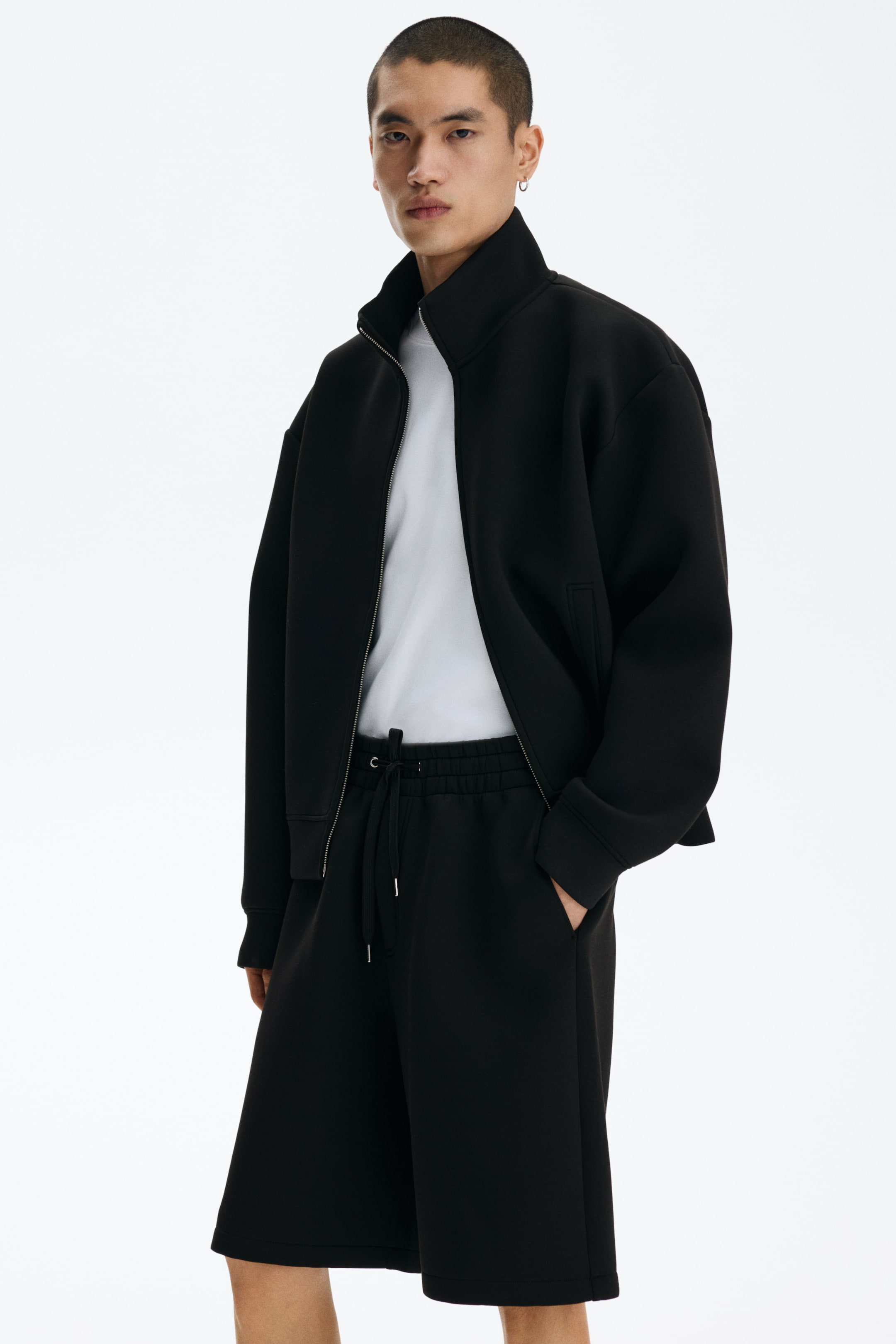 Voir une image plus grande: Une veste noire à fermeture éclair avec un col montant et une fermeture éclair argentée est présentée sous un léger angle, montrant sa coupe décontractée, une poche passepoilée visible et un t-shirt blanc porté en dessous. Cette veste est assortie à un short noir longueur genou, doté d'une taille élastiquée avec cordon de serrage et de poches latérales.