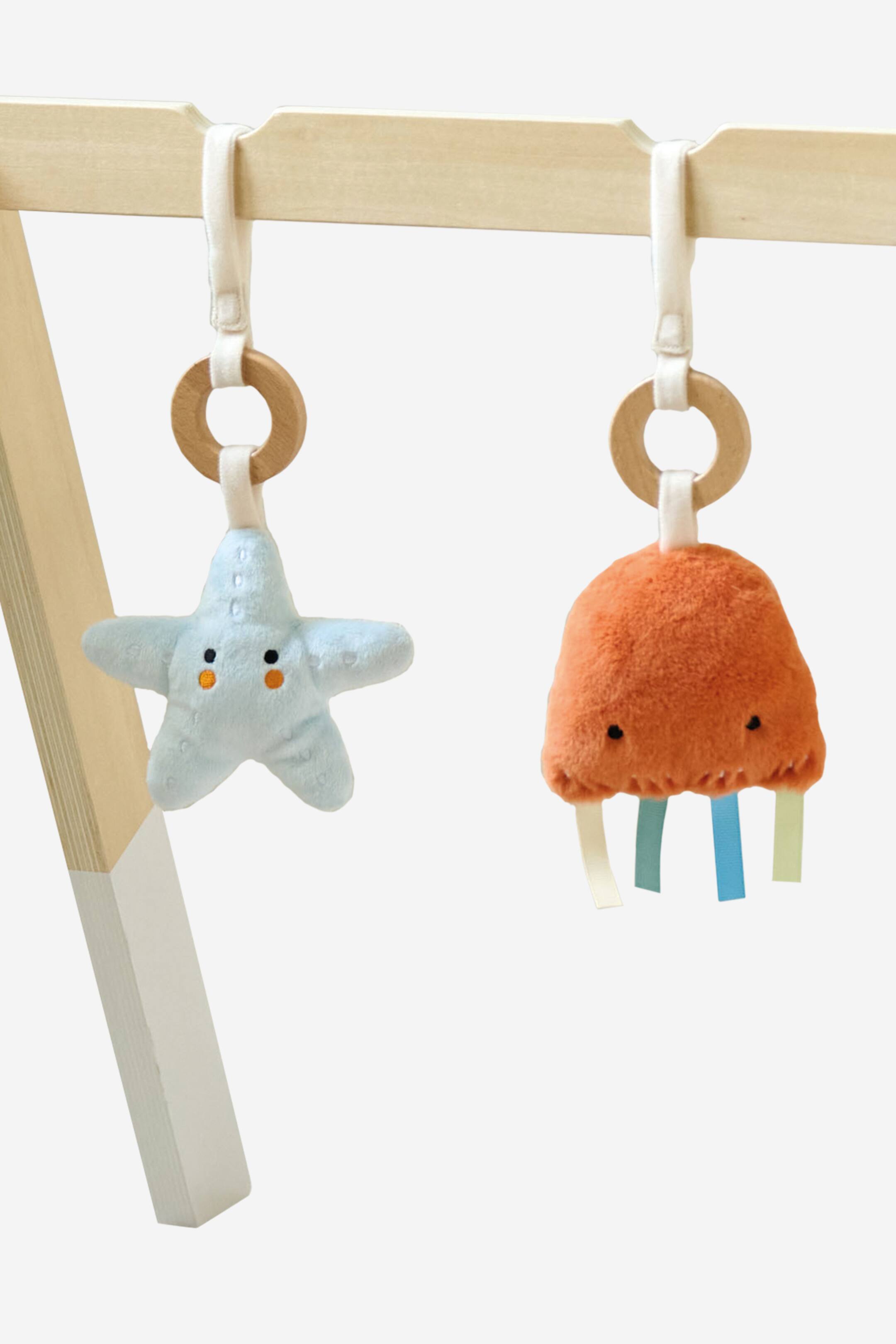 Grotere afbeelding bekijken: Twee pluche babytuigjes hangen aan een houten balk, elk vastgehouden door een witte stoffen lus en een houten ring. Het lichtblauwe, stervormige speelgoed aan de linkerkant heeft geborduurde gezichtsdetails, en het oranje, koepelvormige pluche speelgoed aan de rechterkant vertoont een wollige textuur, geborduurde ogen en kleurrijke stoffen linten die aan de basis bungelen.