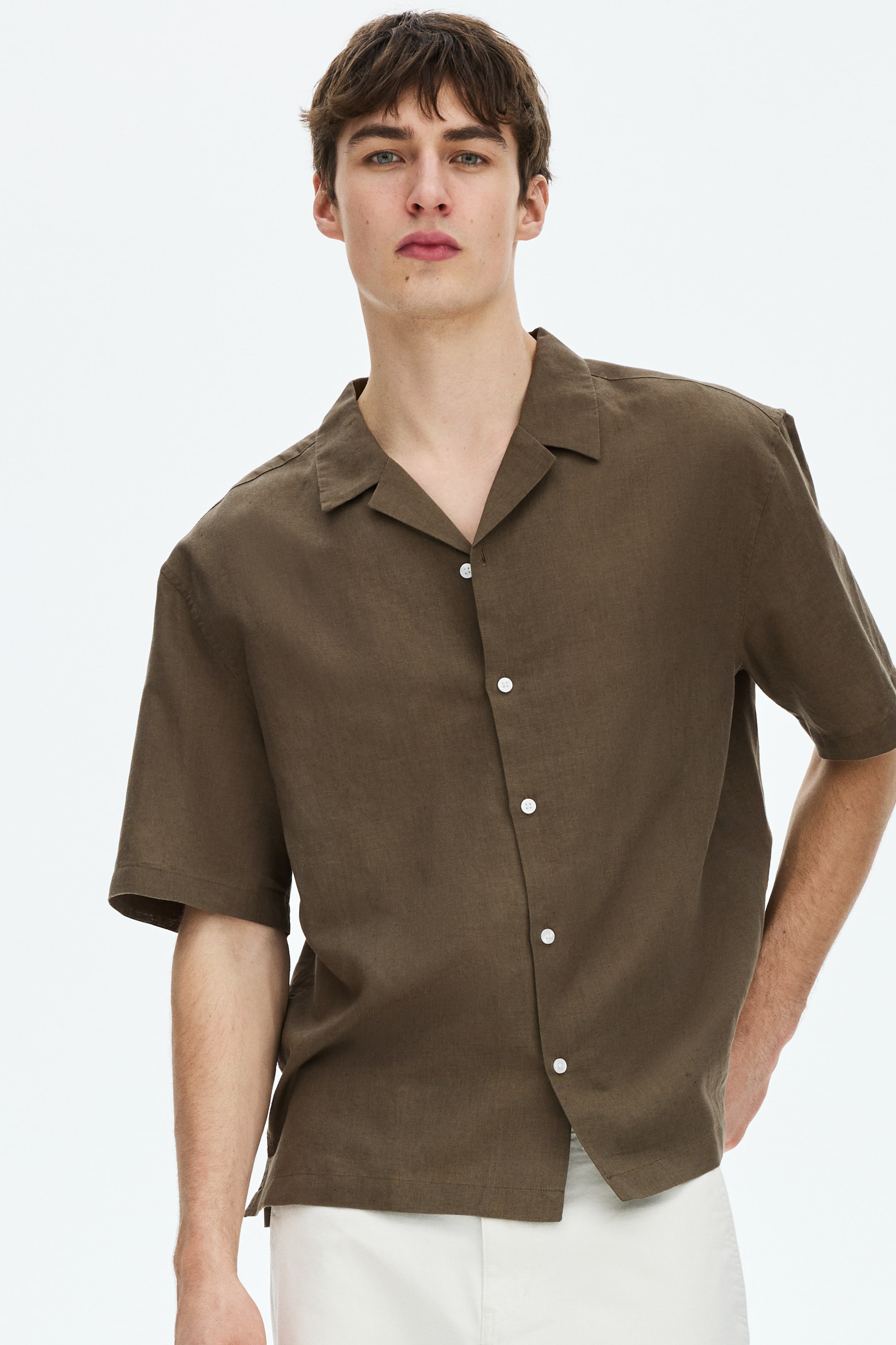 View larger image: A dark olive brown short-sleeved shirt is being worn, featuring a camp collar and a full button-front closure with white buttons. It presents a relaxed fit and a straight hem with side slits.