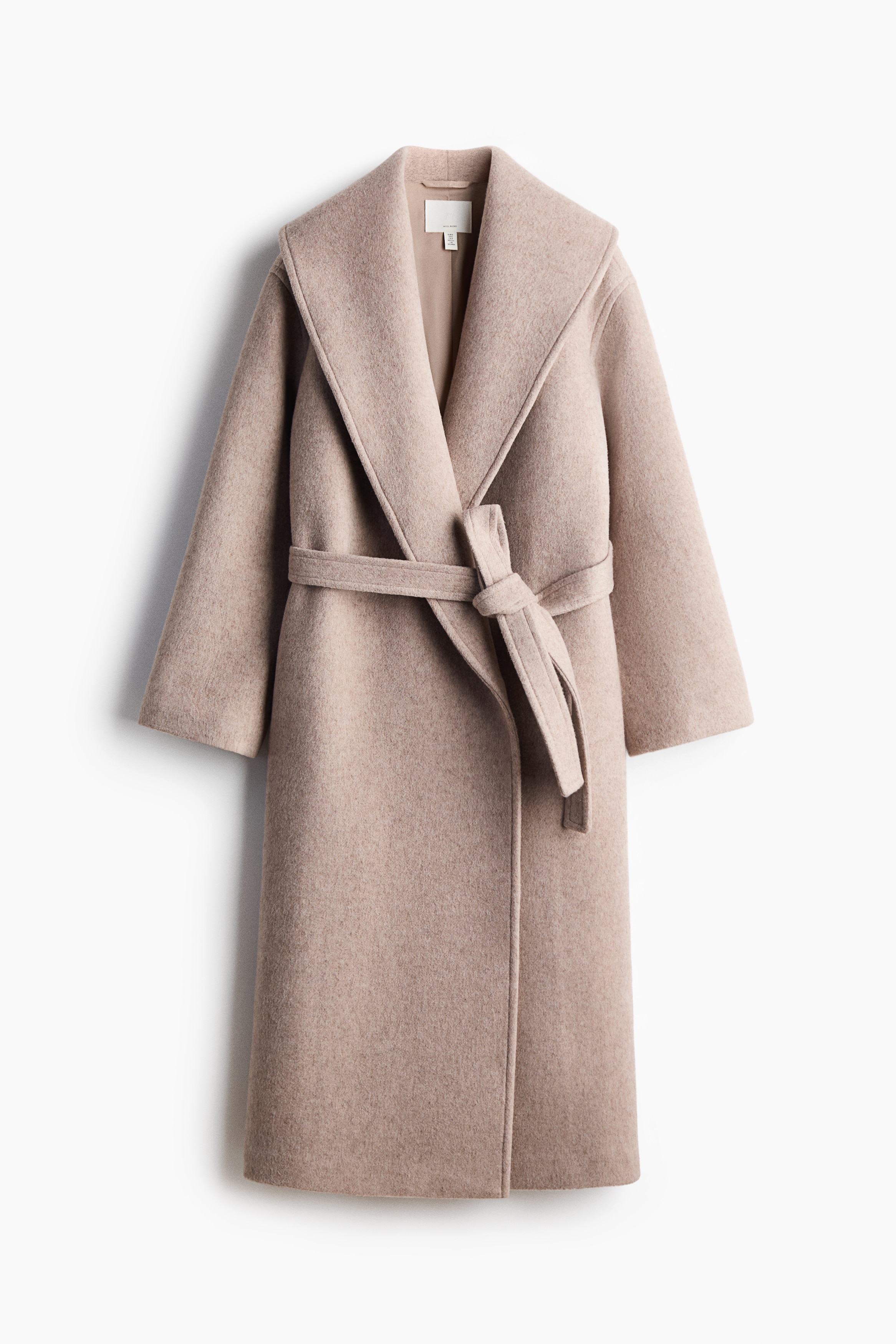 A long, beige felted wool-blend coat is shown from the front, featuring a wide shawl collar and a matching tie belt knotted at the waist. The relaxed-fit coat is styled with hands placed in its discreet side pockets, with white cuffs peeking out from the sleeves.