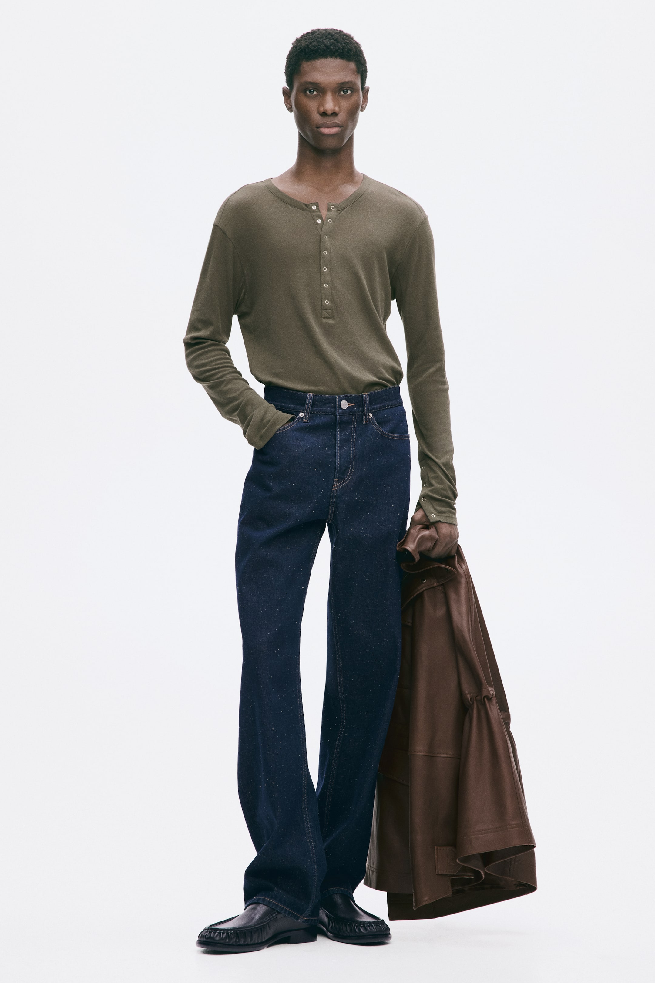 Büyük resmi görüntüle: Modelin üzerinde, düğmeli patletli zeytin yeşili uzun kollu bir henley gömlek var ve koyu yıkamalı geniş paça jean pantolonun içine sokulmuş. Model ayrıca siyah moc-toe loafer ayakkabılar giyiyor ve kahverengi deri ceketini kolundan taşıyor.