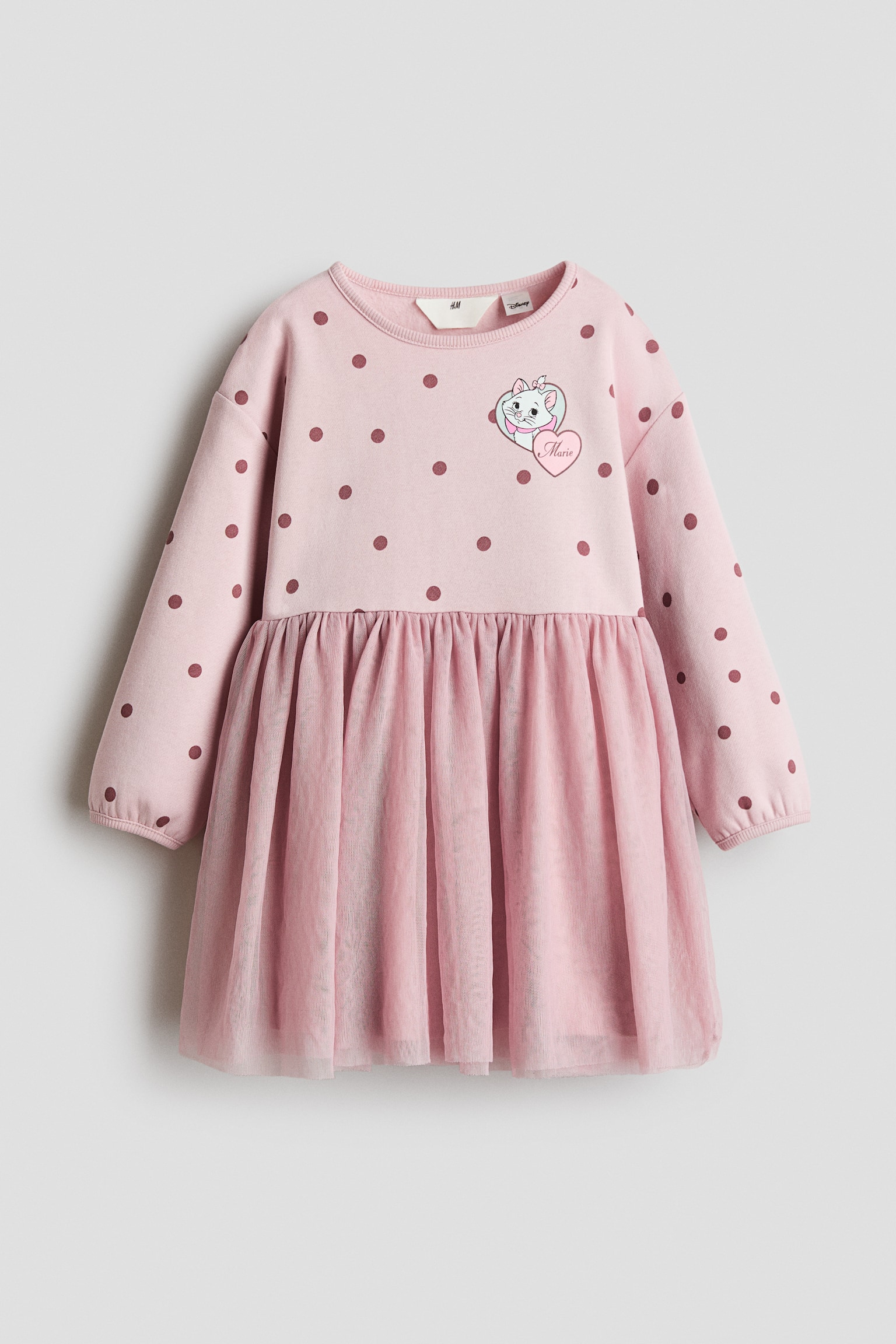 A light pink long-sleeved dress is shown from the front, featuring a polka-dot patterned bodice and a gathered tulle skirt. The bodice features a ribbed crew neck, long sleeves with elasticated cuffs, and a small heart-shaped graphic of a white cat on the left chest. The matching pink tulle skirt falls from the gathered waist seam in a flared silhouette.