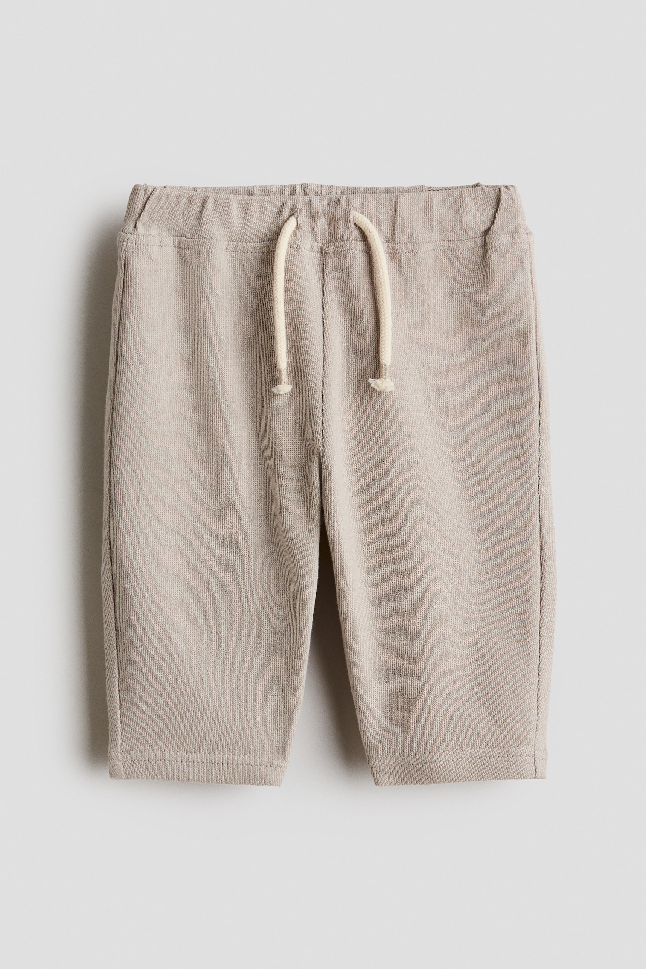 Grösseres Bild ansehen: Die Hosen werden von vorne präsentiert und zeigen ein weiches, hellbeiges, strukturiertes Jersey-Material. Sie verfügen über einen verstellbaren, elastischen Bund und eine dekorative, cremefarbene Kordel mit geknoteten Enden.