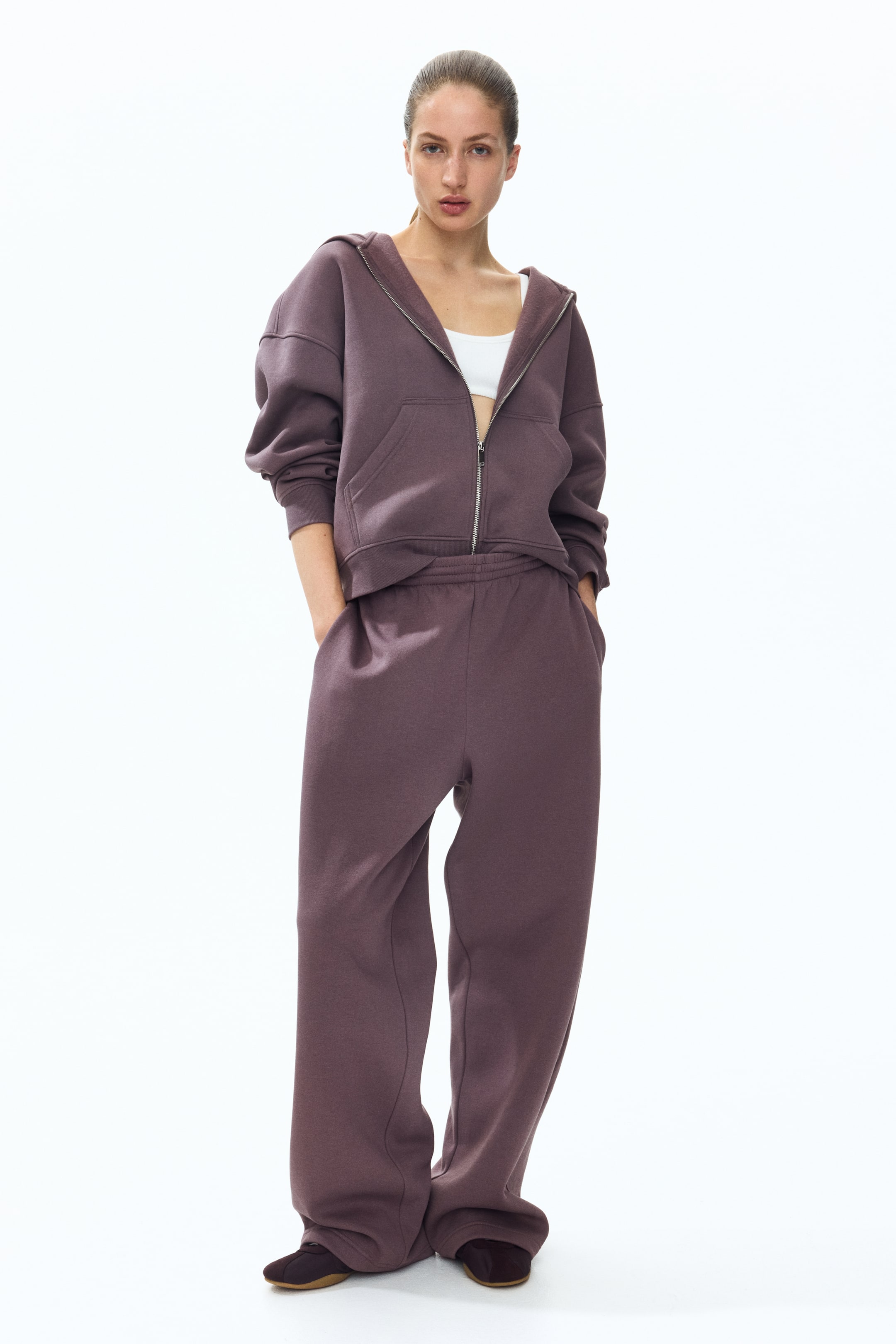 View larger image: The model is wearing dark plum-colored sweatpants made from sweatshirt fabric, paired with a matching hooded sweatshirt. The sweatpants have a smocked elastic waistband, side pockets where the model's hands are resting, and straight legs.