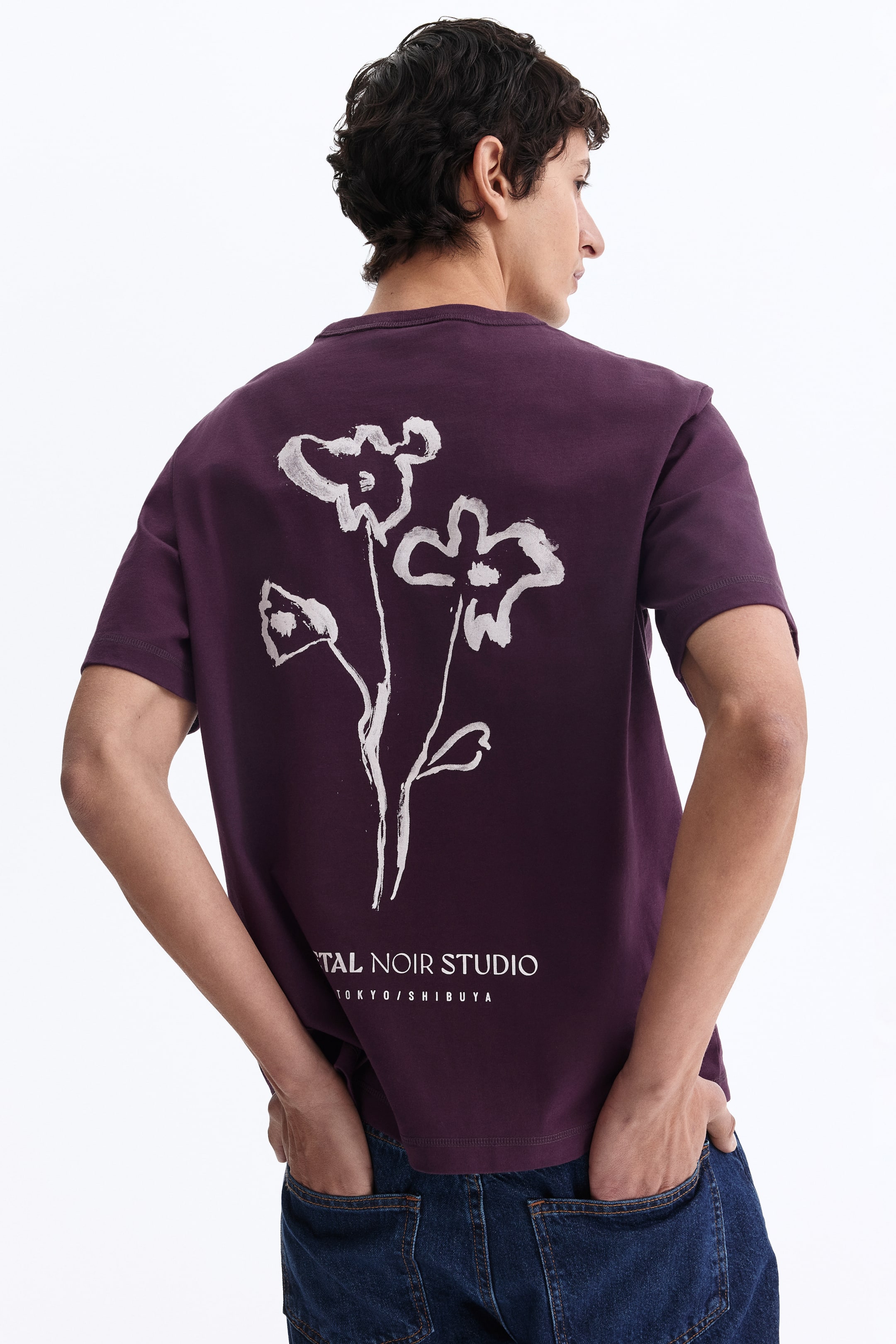 View larger image: A deep-purple short-sleeved T-shirt features a white, stylised floral graphic on the back. Below the graphic, white text reads 'TAL NOIR STUDIO TOKYO/SHIBUYA'. Seen from the back, the T-shirt shows a relaxed fit.