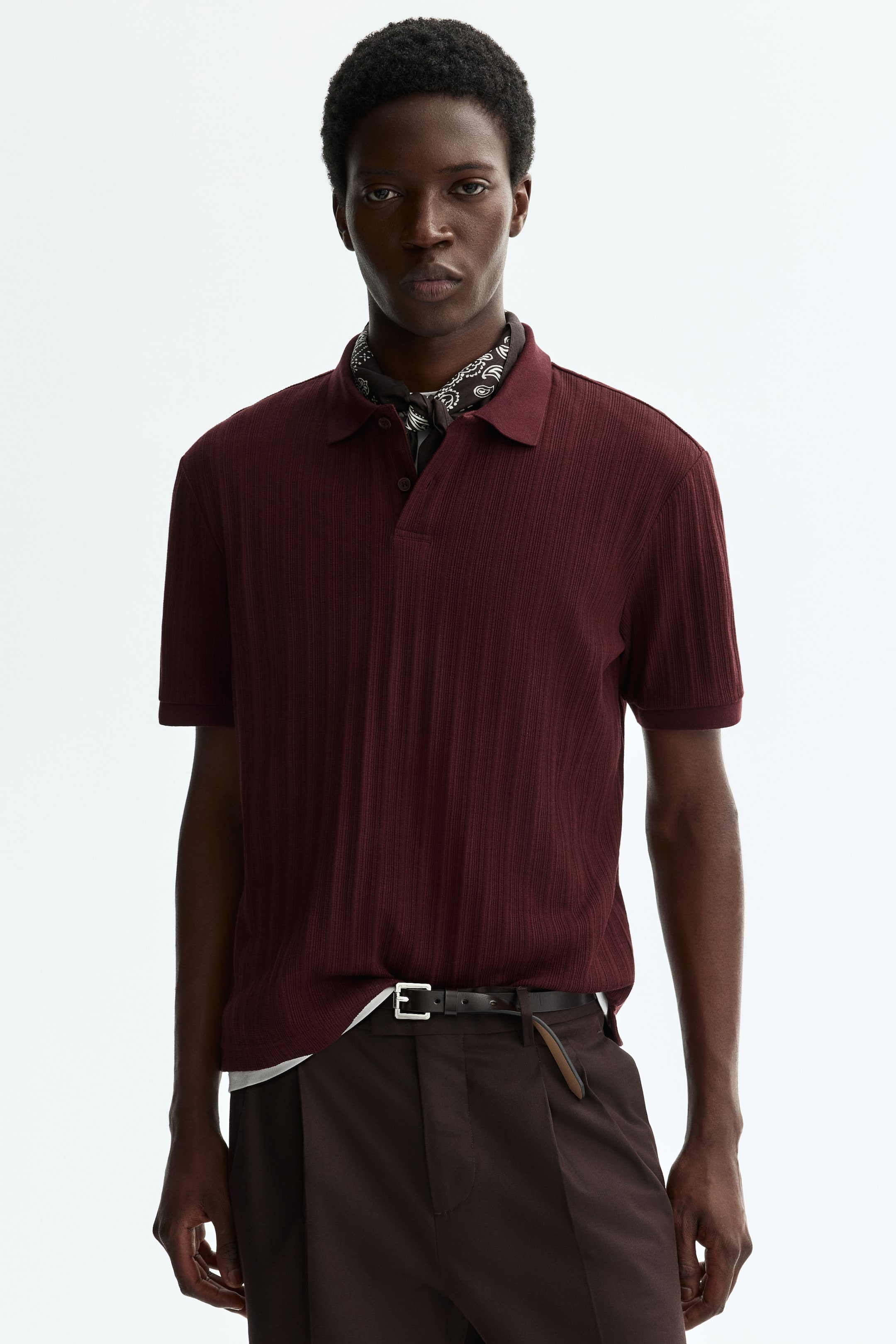 Grösseres Bild ansehen: Ein dunkelrotes, geripptes Poloshirt ist nach vorne gerichtet und verfügt über einen Polokragen, eine Zwei-Knopf-Leiste und kurze Ärmel mit gerippten Bündchen. Ein schwarz-weiß gemustertes Bandana ist unter dem Kragen sichtbar, und ein weißes Unterhemd ist am Saum zu sehen.