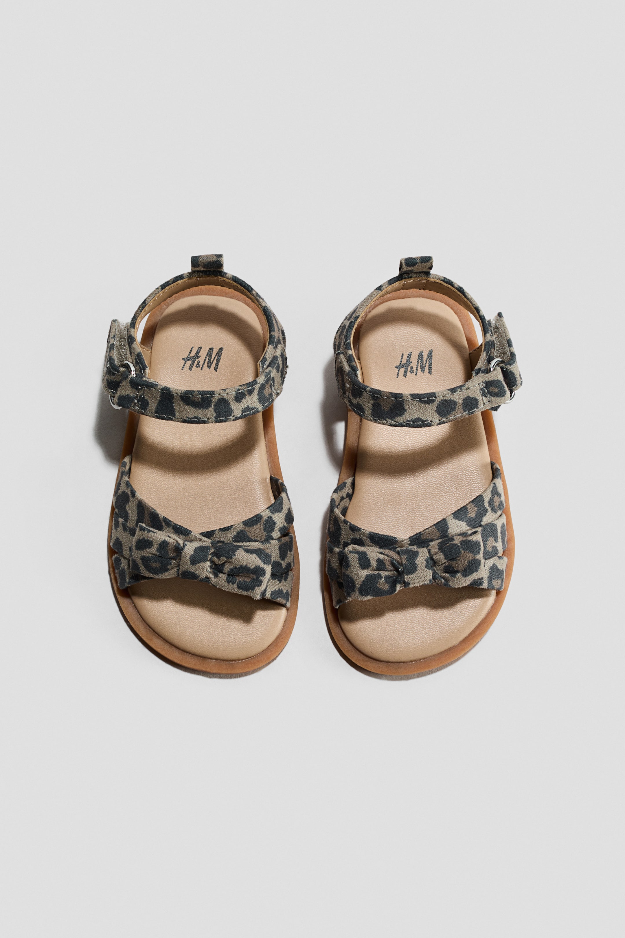 Ver imagem maior: Um par de sandálias com estampa de leopardo em tons de bege e preto é apresentado numa vista de cima. Cada sandália possui uma palmilha em bege claro com o logótipo da H&M e uma sola castanha. Uma tira larga sobre o peito do pé exibe um detalhe de laço, e a tira no tornozelo é fixada com um fecho de velcro.
