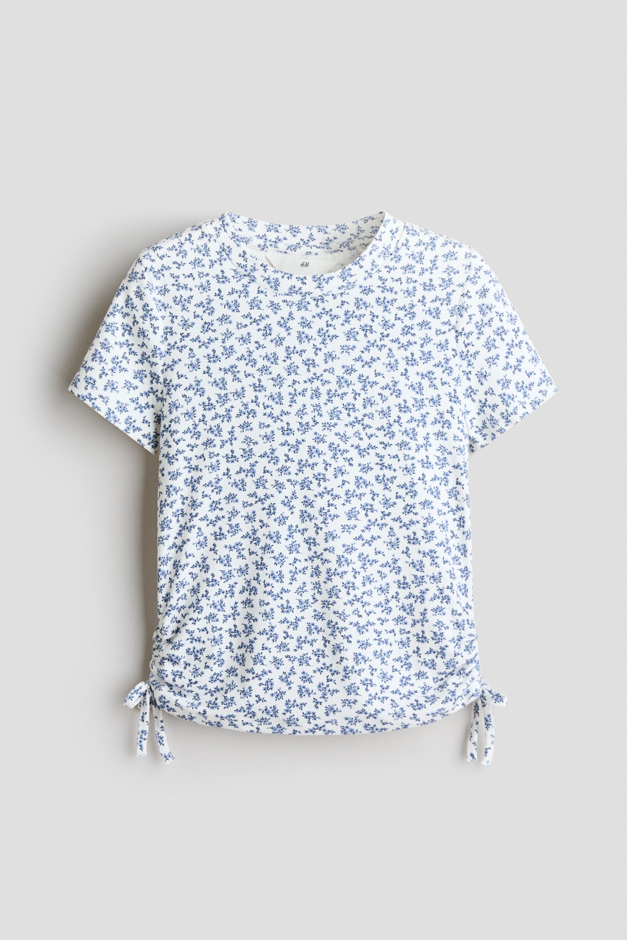 Grotere afbeelding bekijken: Een kort mouwtopje in zachte katoenen jersey is frontaal zichtbaar, met een witte basis en een kleine blauwe bloemenprint. Het heeft een ronde halslijn en decoratieve trekkoorden die aan elke kant van de zoom in strikjes zijn gebonden, waardoor er plooien ontstaan.