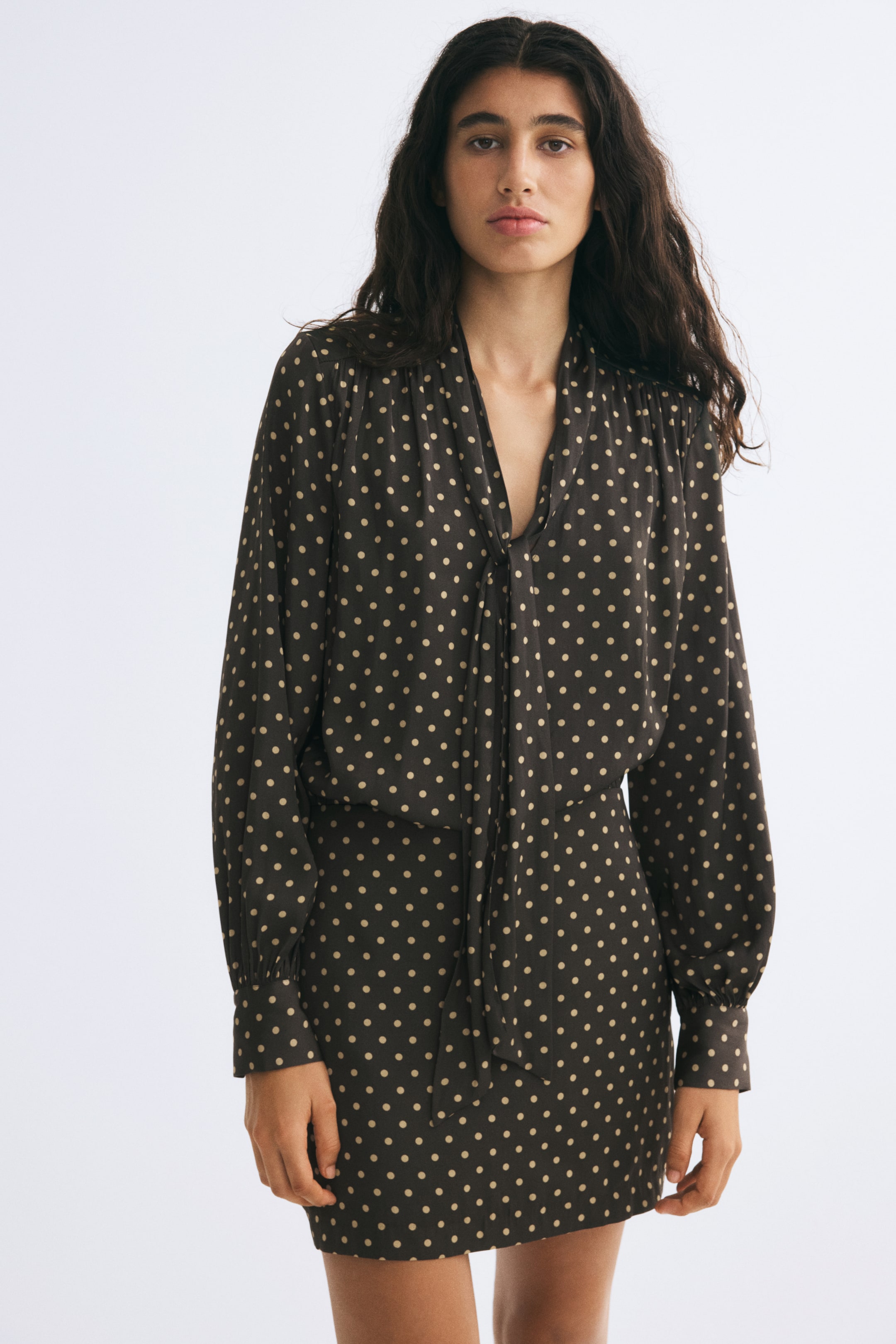 View larger image: A short, polka-dot dress in a dark woven fabric is being worn by the model. The dress features a V-neckline with a tie-up collar, gathered shoulders, and long sleeves with wide button cuffs.