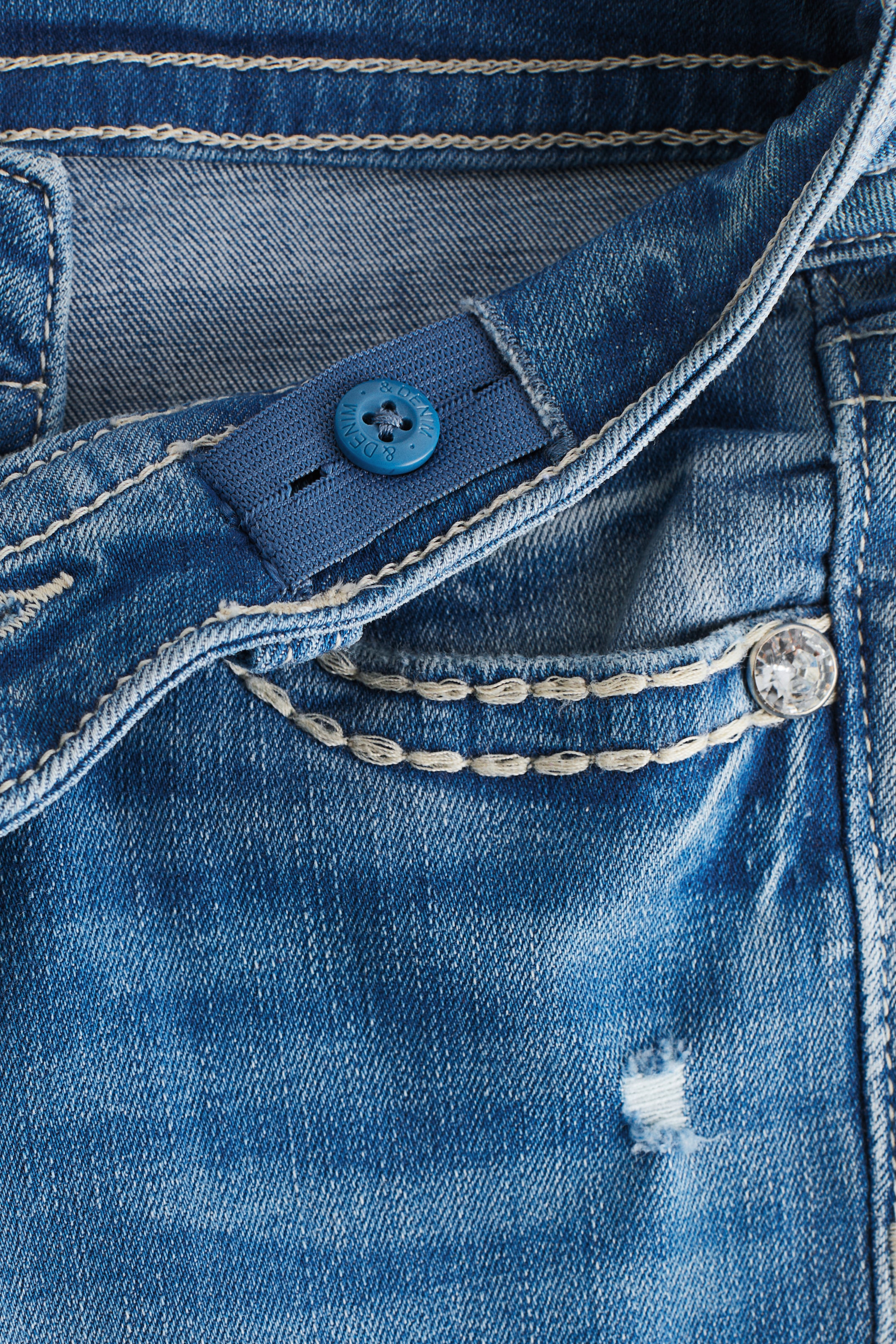 Agrandir l'image: Le jean en denim bleu est présenté en vue de face, avec une ceinture élastique ajustable ornée d'un bouton bleu. Une double couture de couleur claire met en valeur une petite poche agrémentée d'un bouton transparent facetté, et une déchirure usée est visible sur le denim.