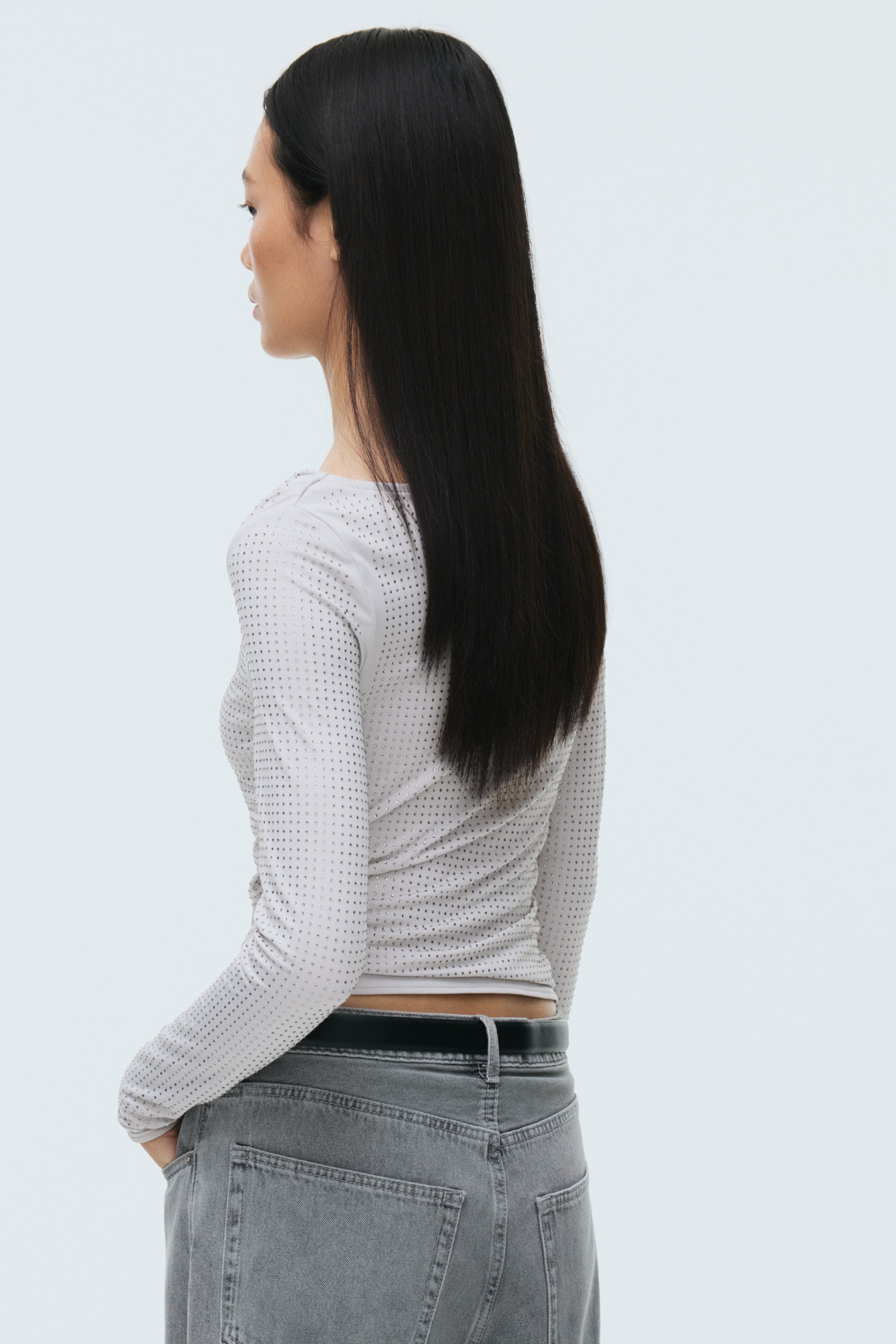 Grösseres Bild ansehen: Ein hellgraues Langarm-T-Shirt wird aus einem seitlich-hinteren Winkel präsentiert. Es zeigt eine körperbetonte Silhouette mit kleinen, rundum angebrachten Nieten und wird mit grauen Jeans im Used-Look und einem schwarzen Gürtel gestylt.
